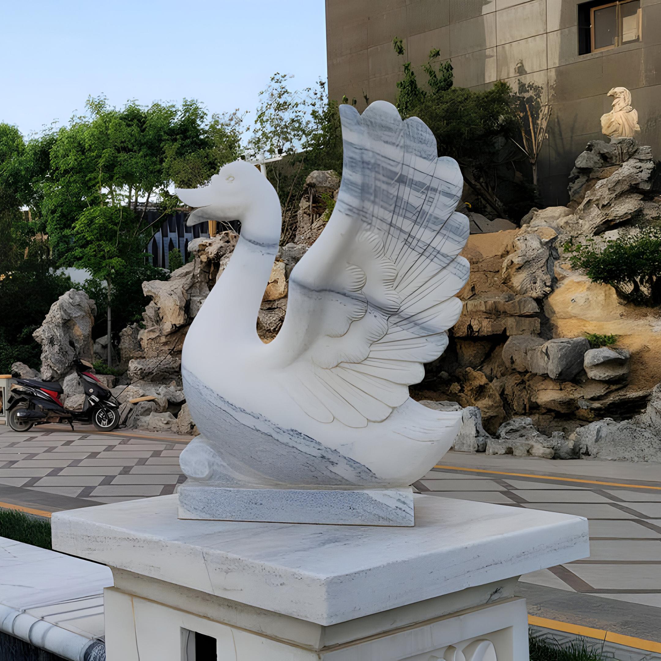 The Wings-Lifted Swan I Marble Outdoor Sculpture by Giant Sculptures, standing at 120cm and meticulously hand-carved, gracefully stands on a pedestal with outstretched wings. It is surrounded by rocky landscaping, lush greenery, a building facade, and a parked motorbike.
