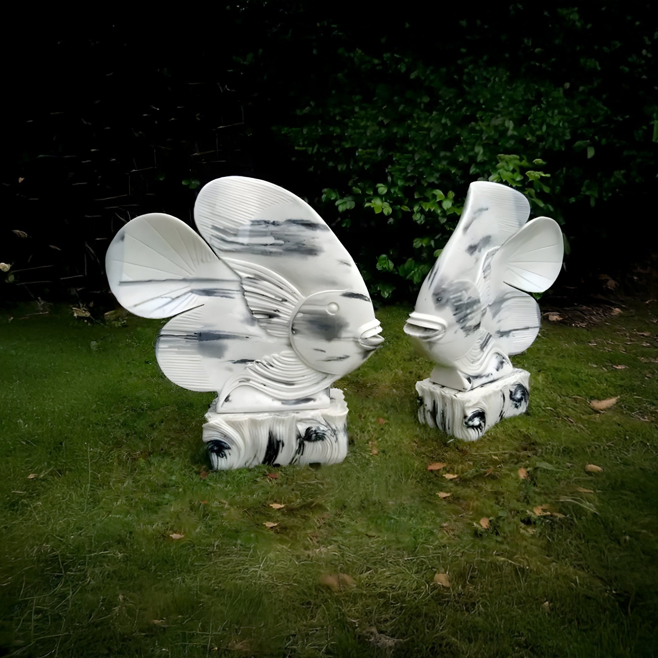 Two Swirl-Fin Angel Fish II Marble Outdoor Sculptures by Giant Sculptures stand on grass, white with black marbling and exaggerated fins and eyes. Lush green foliage creates a serene setting for this enchanting marine-inspired display.