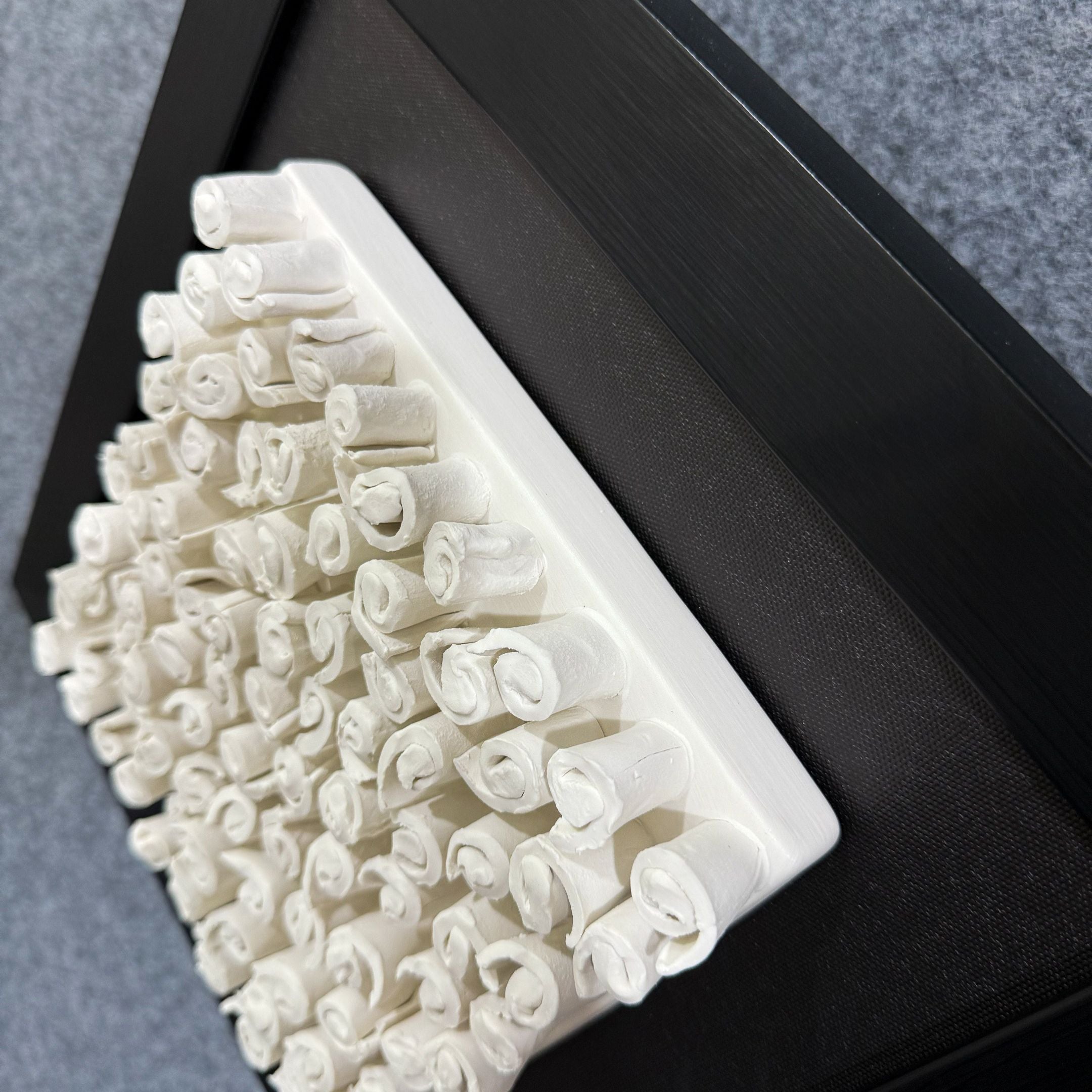 The Harmonia 3D Spiral Wall Sculpture by Giant Sculptures features numerous upright white paper rolls in rows within a rectangular base and black frame, ideal for adding contemporary flair to interiors against a gray backdrop.
