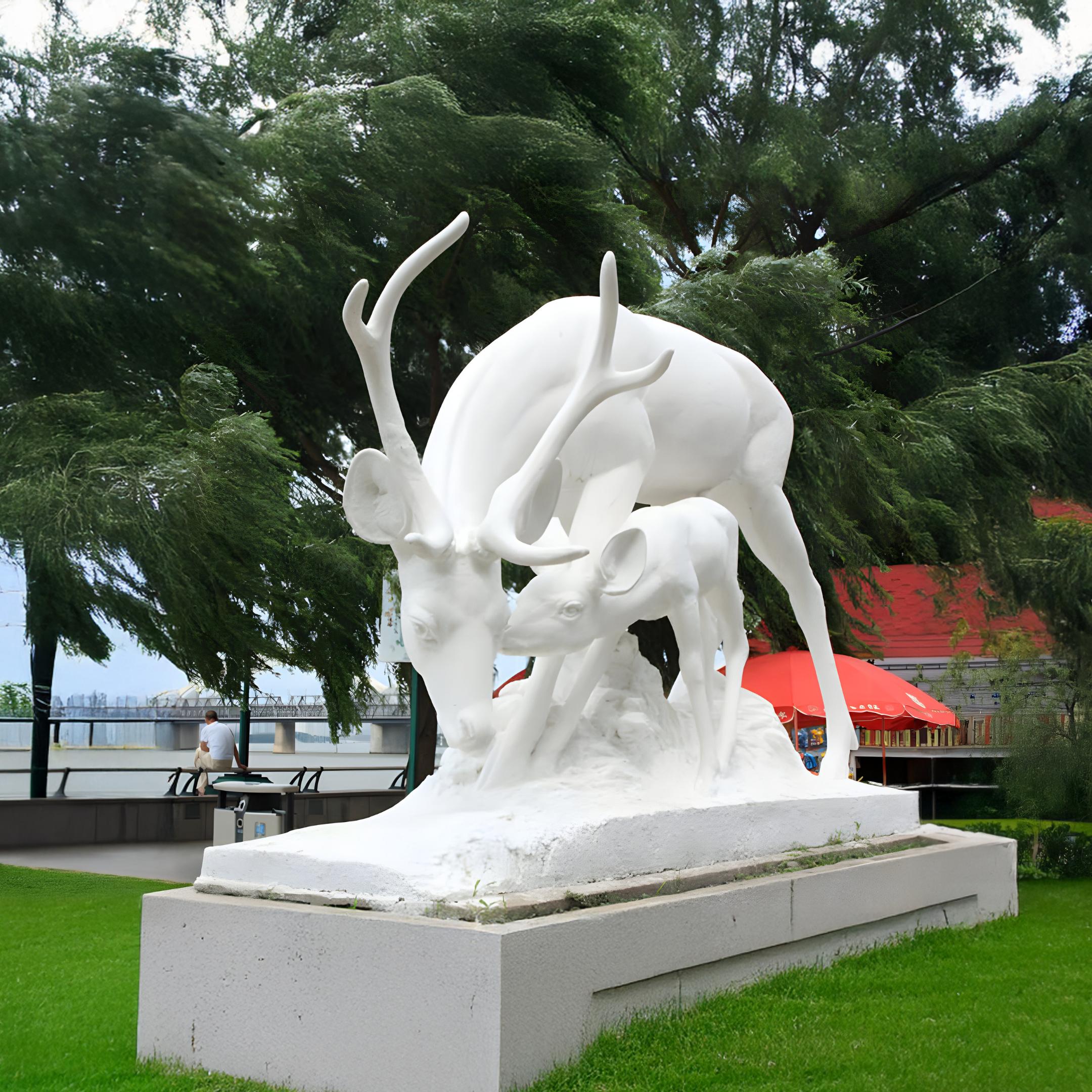 The Nurturing Deer Marble Outdoor Sculpture - 160cm by Giant Sculptures graces the park, set on a platform amid lush grass and swaying trees. This exquisite piece of garden art enhances the view of the red-roofed structure against a cloudy sky backdrop.