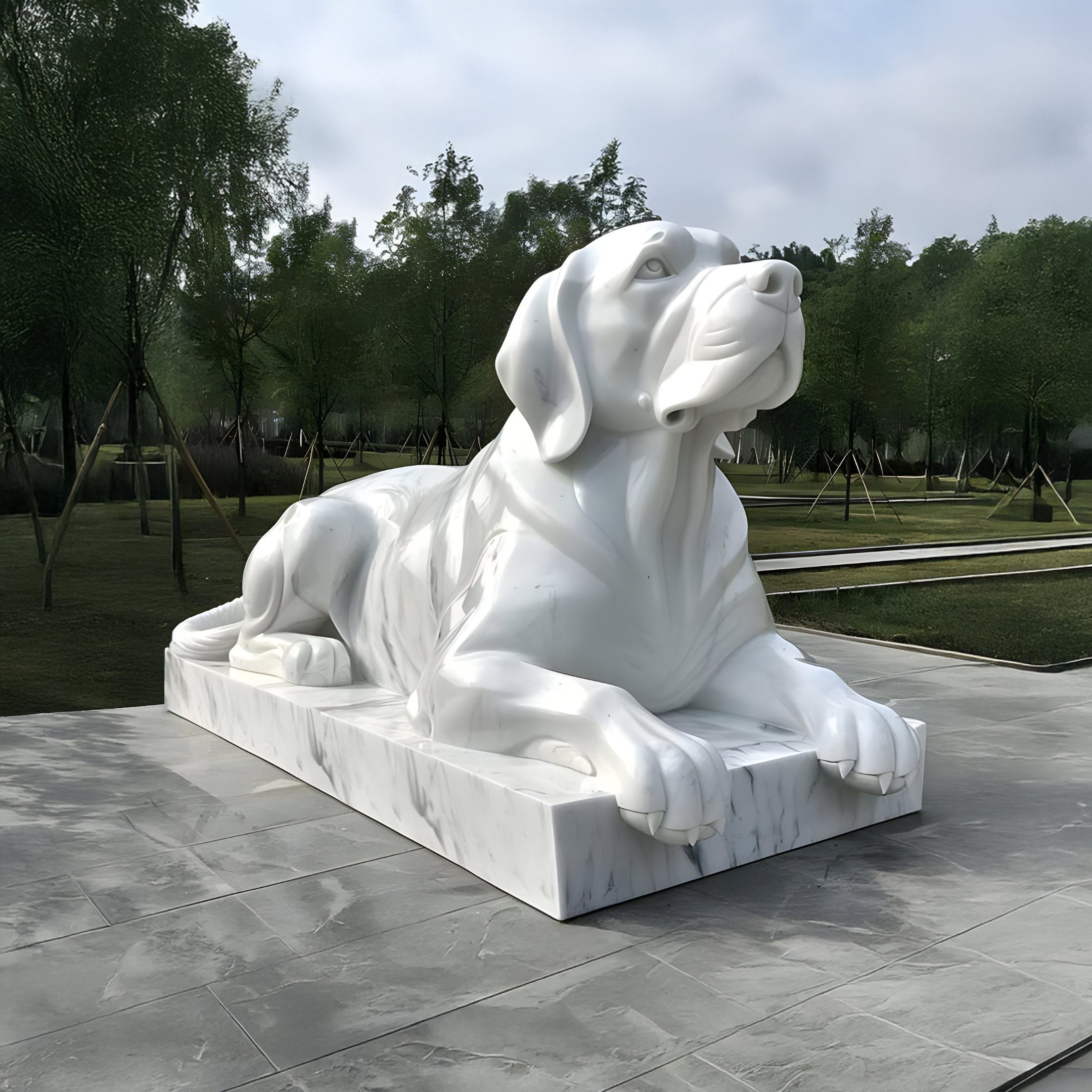 The park features the Attentive Lying Dog Marble Outdoor Sculpture (90cm) by Giant Sculptures on a rectangular platform. The neatly arranged trees and cloudy sky create a serene backdrop, perfect for appreciating this outdoor artwork.
