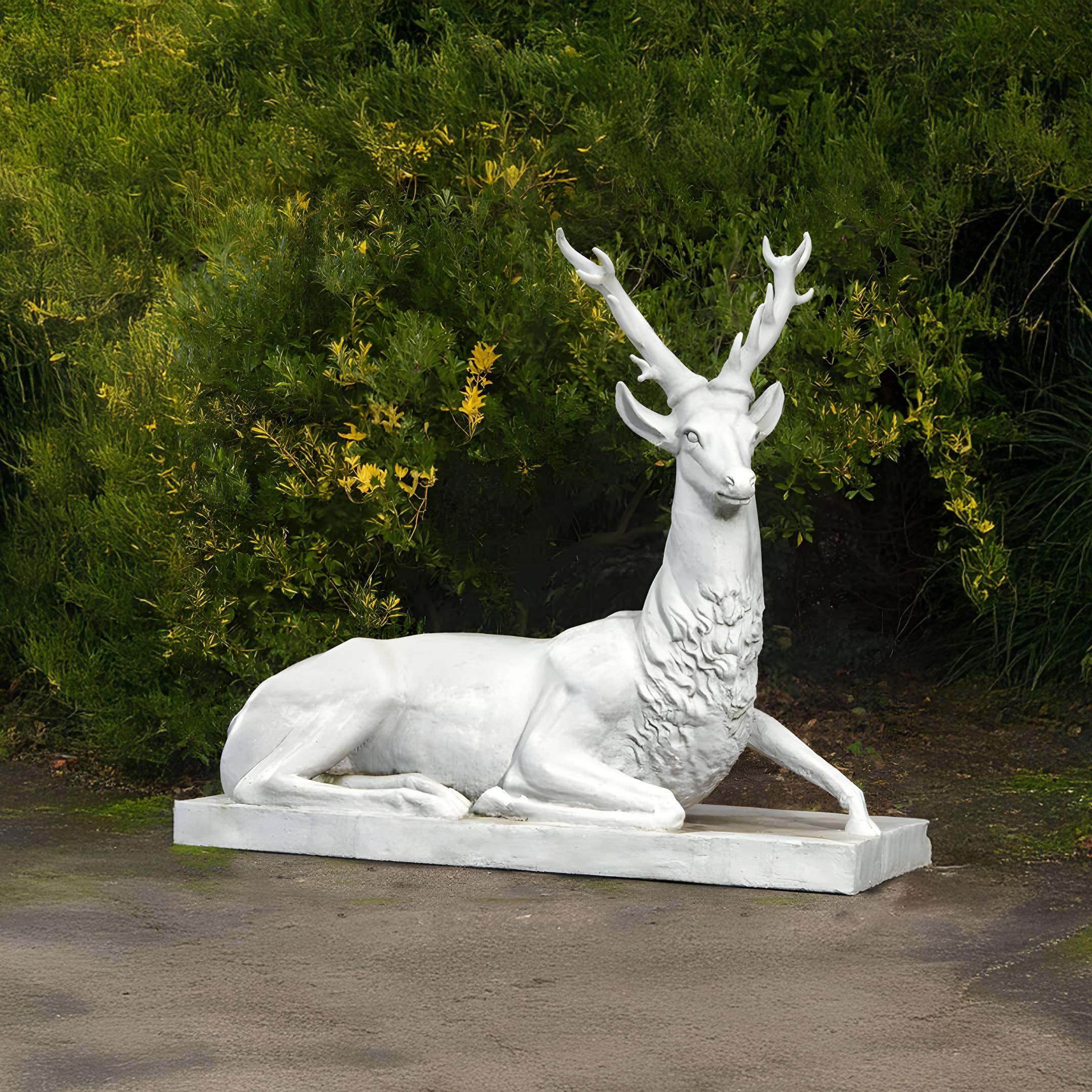The Regal Lying Stag Marble Outdoor Sculpture by Giant Sculptures, a 150cm handcrafted piece, features a graceful white deer with large antlers reclining on a rectangular base. It elegantly adorns paved areas, surrounded by lush green foliage.