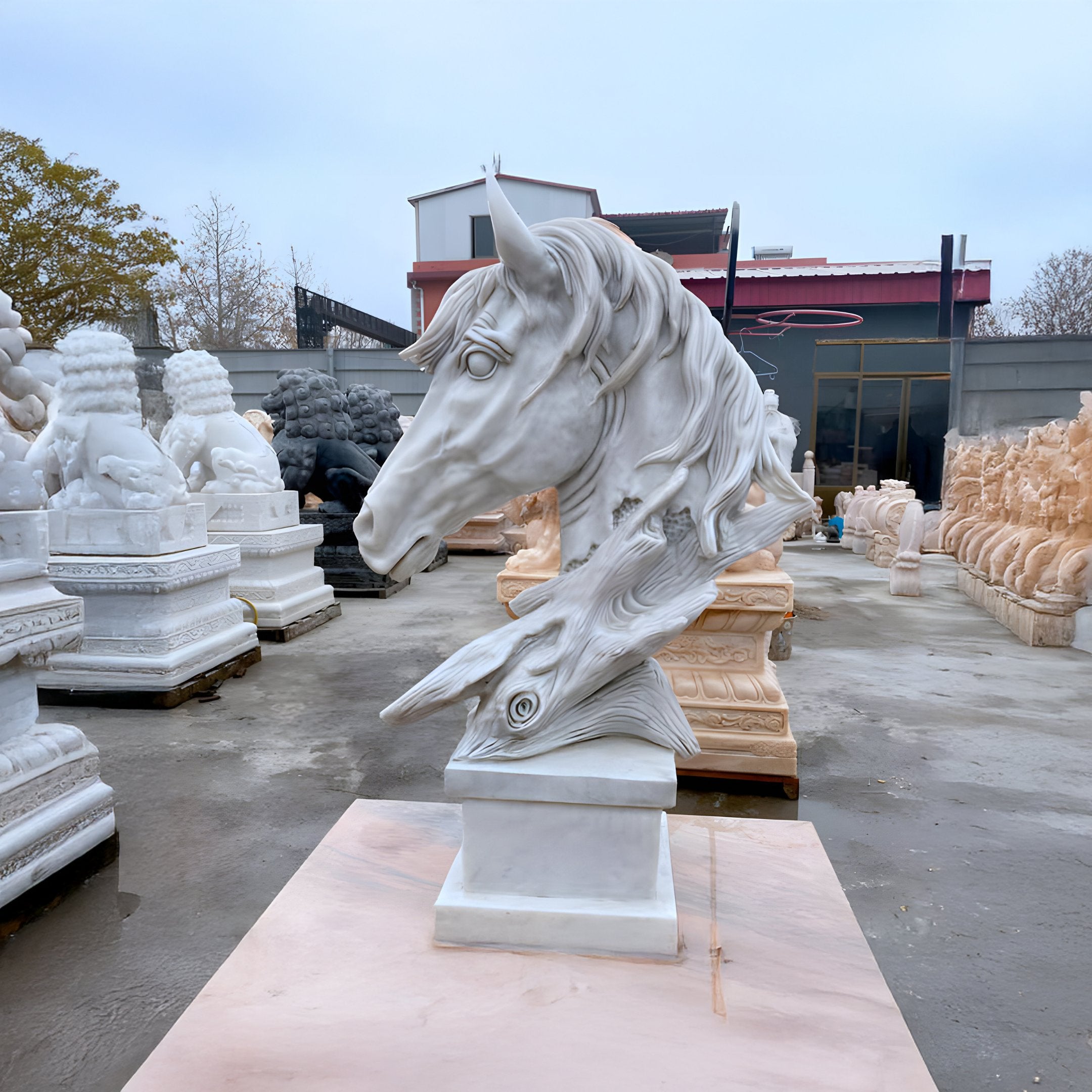 The 100cm Dynamic Horse Head Marble Outdoor Sculpture by Giant Sculptures gracefully sits on a pedestal amidst other yard sculptures. Its detailed flowing mane and expressive eyes form an elegant scene, complemented by surrounding buildings and leafless trees.