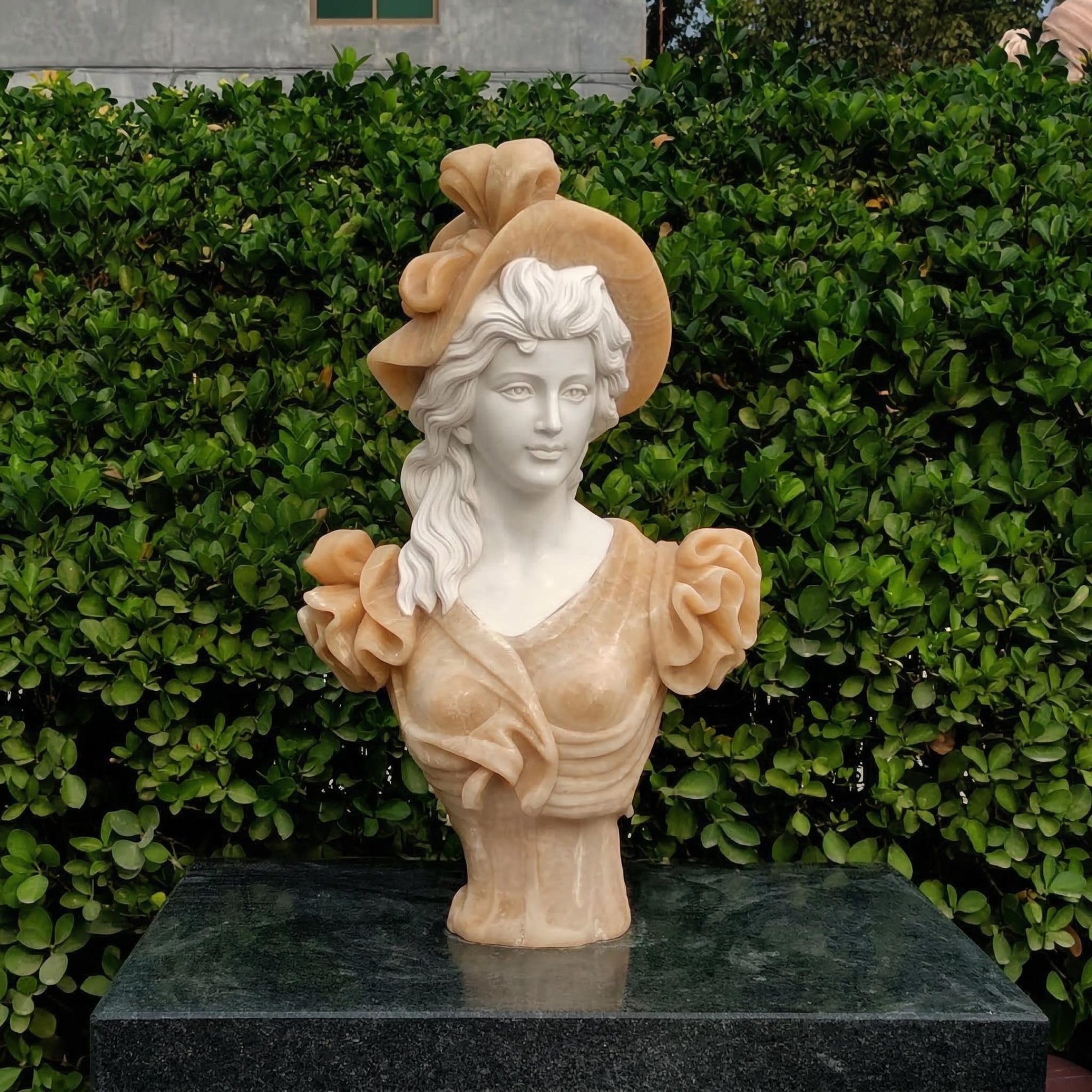 The Classical Lady Marble Bust - 65cm by Giant Sculptures, featuring wavy hair, a hat with a large bow, and a ruffled dress, is shown on a dark stone pedestal in front of dense green bushes.