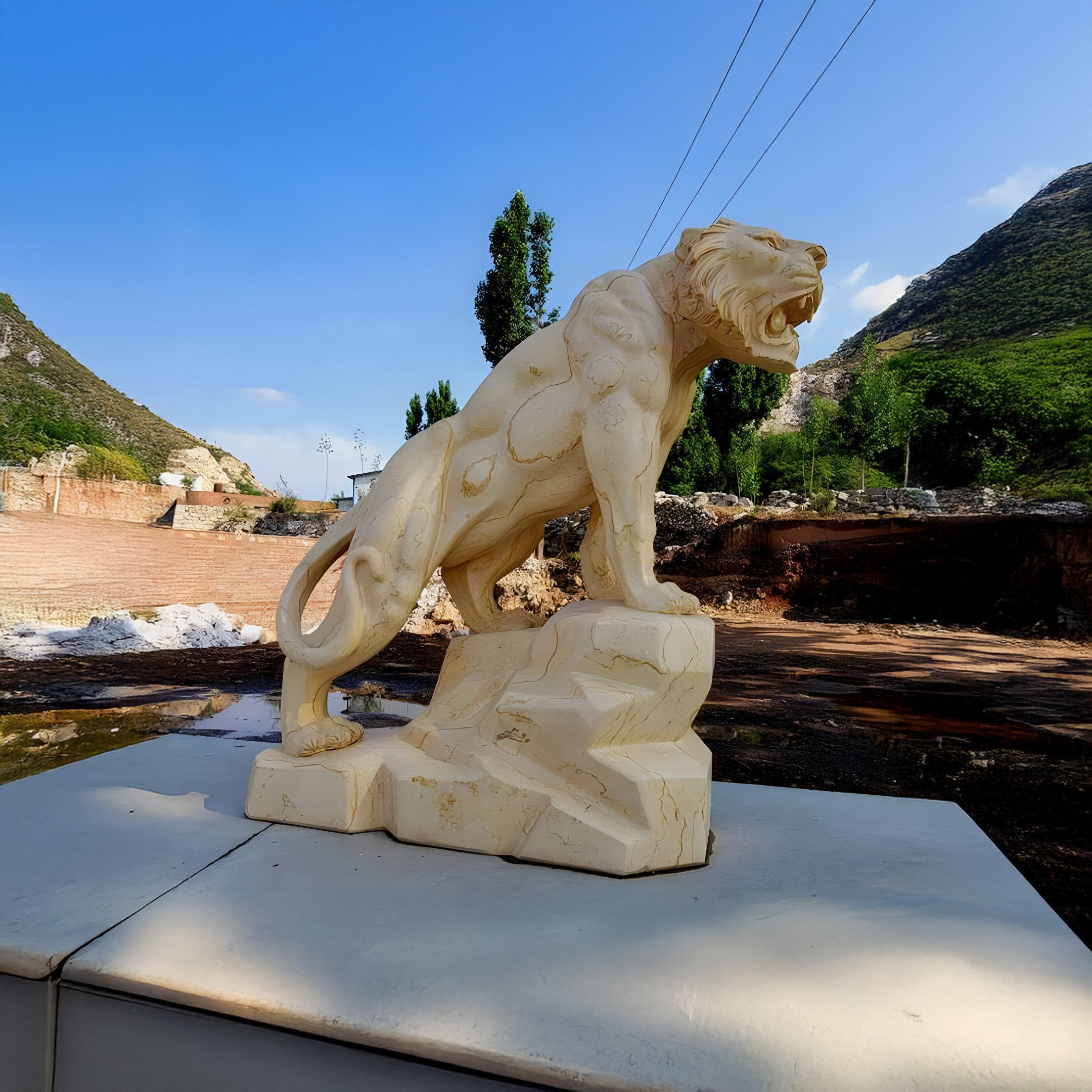 The Roaring Rock Tiger Yellow Marble Outdoor Sculpture by Giant Sculptures features a cream-colored stone lion on a pedestal, roaring and facing left with mountains and a clear blue sky in the background.