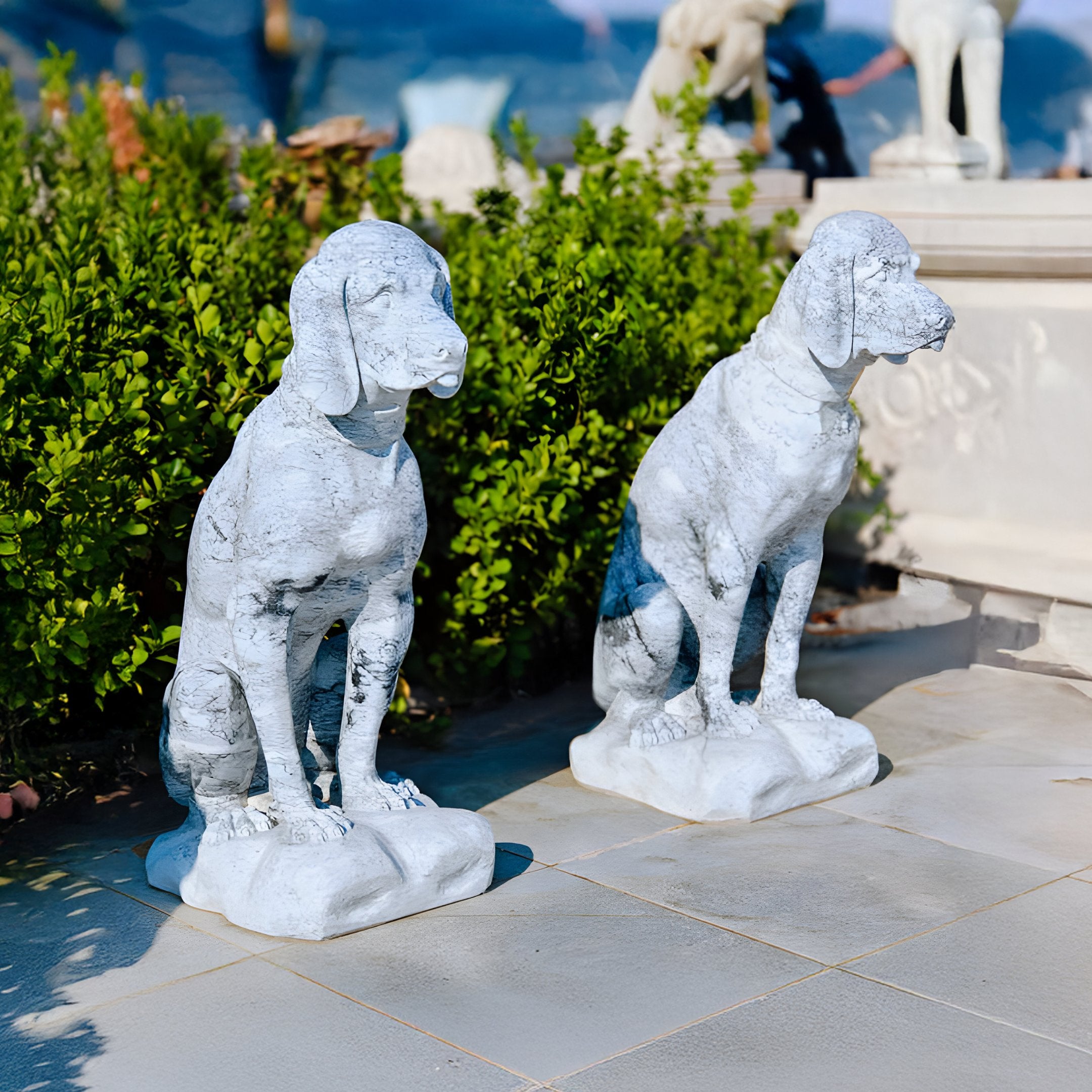 Two Watchful Hound Dog Marble Outdoor Sculptures by Giant Sculptures, each 100cm tall, grace a tiled pathway framed by lush green bushes. Their textured surfaces complement a blurred sculpture in the background beneath a clear blue sky.
