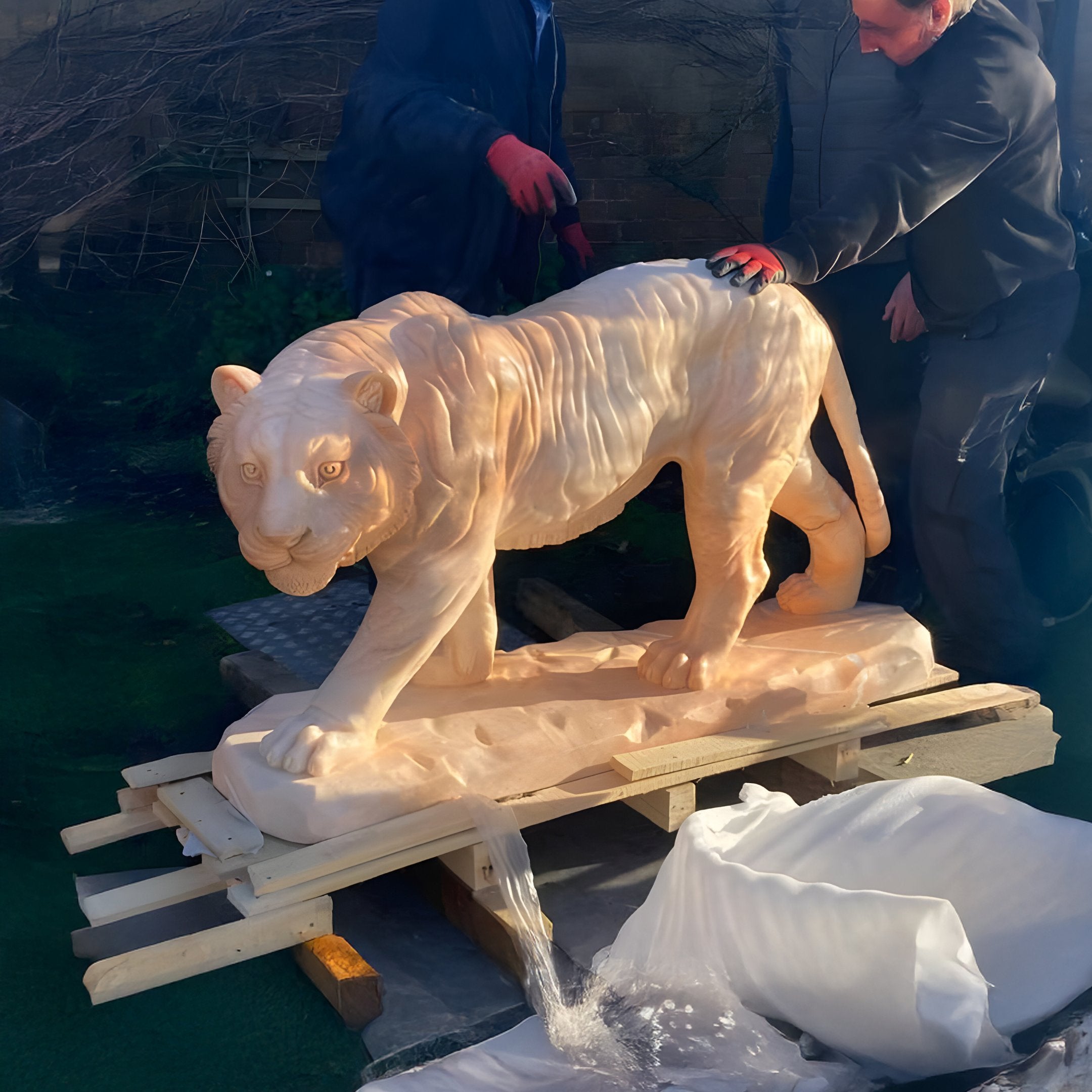 Two people wearing gloves carefully uncover a life-sized Striding Tiger Yellow Marble Outdoor Sculpture by Giant Sculptures. The 60cm piece, rich with intricate details, rests on a wooden pallet surrounded by packaging material, seamlessly blending art with nature.