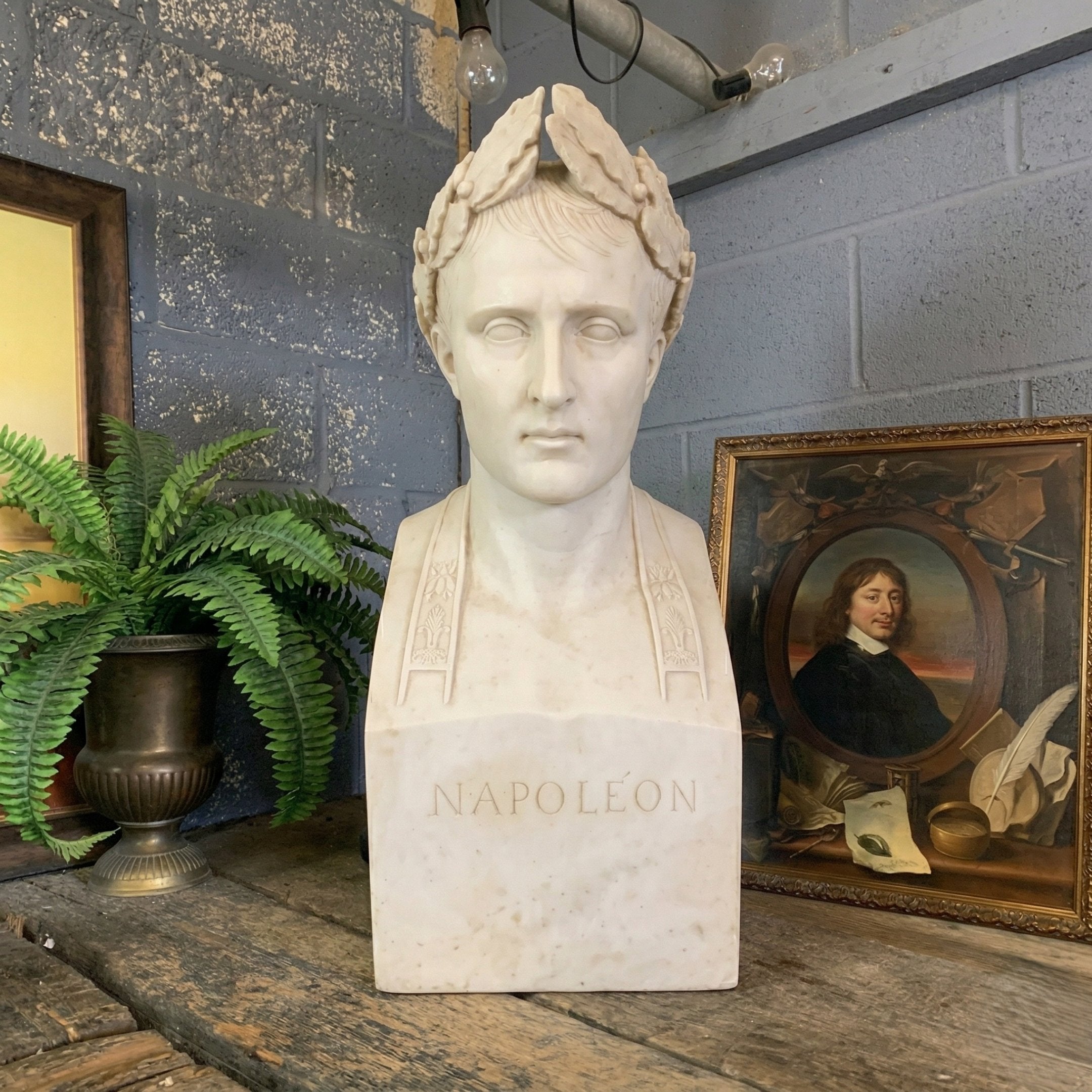 The Napoleon Marble Bust Sculpture - 60cm by Giant Sculptures rests on a wooden table between a potted fern and a framed portrait, adding historic charm and elegance to the scene against the gray brick wall.