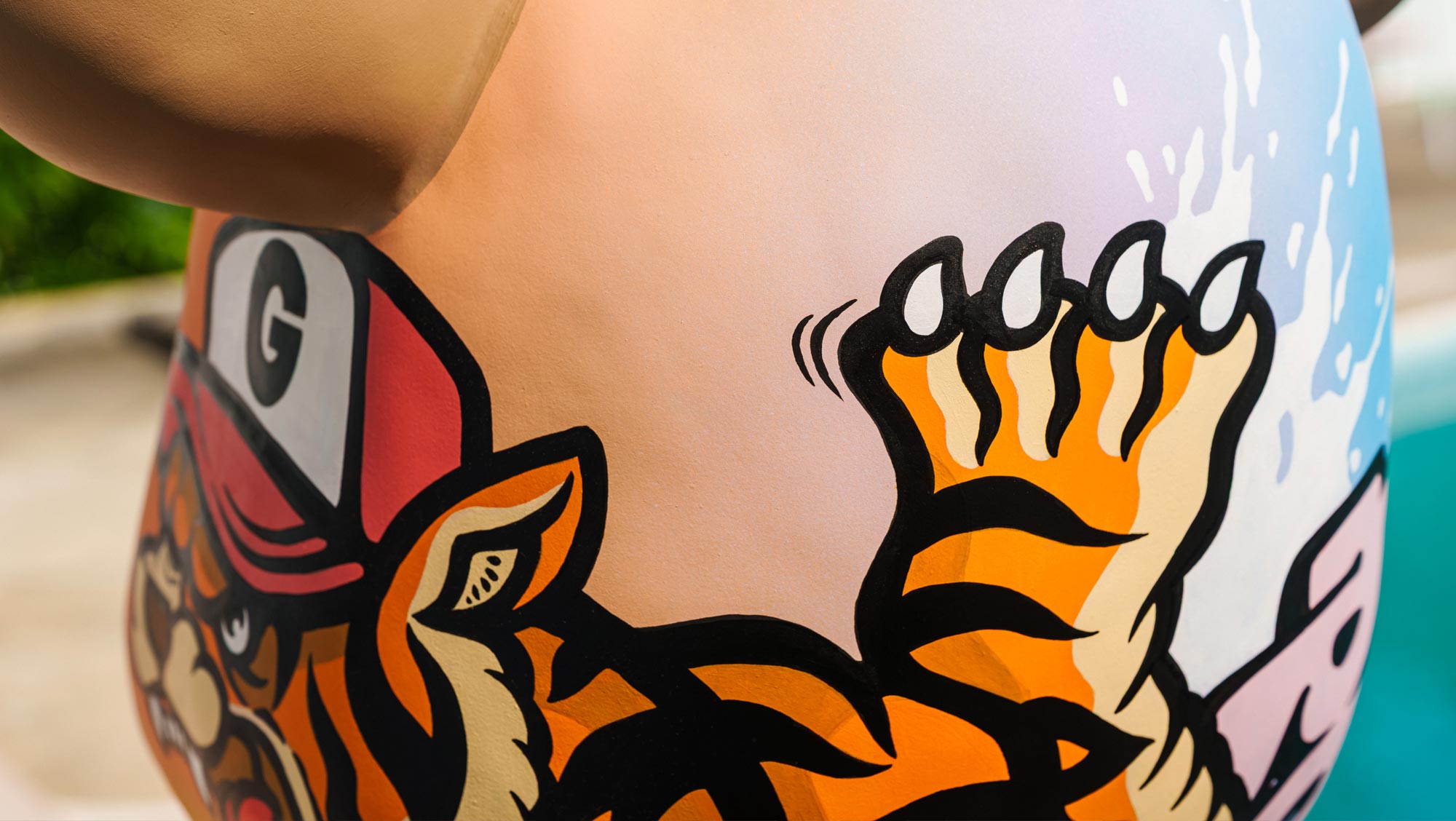 Close-up of a cartoon tiger wearing a red and white cap, with one orange and black striped paw raised, painted on a rounded surface next to a blue area that resembles water.