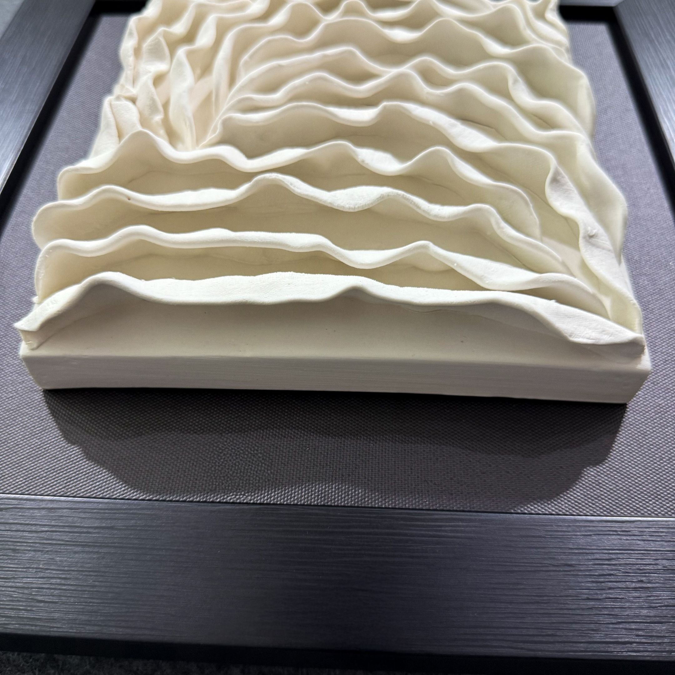 The Harmonia 3D Ceramic Wave Wall Sculpture by Giant Sculptures features wavy white ceramic layers in a black frame, creating a striking rippled effect—a unique textured wall installation perfect for modern interiors.