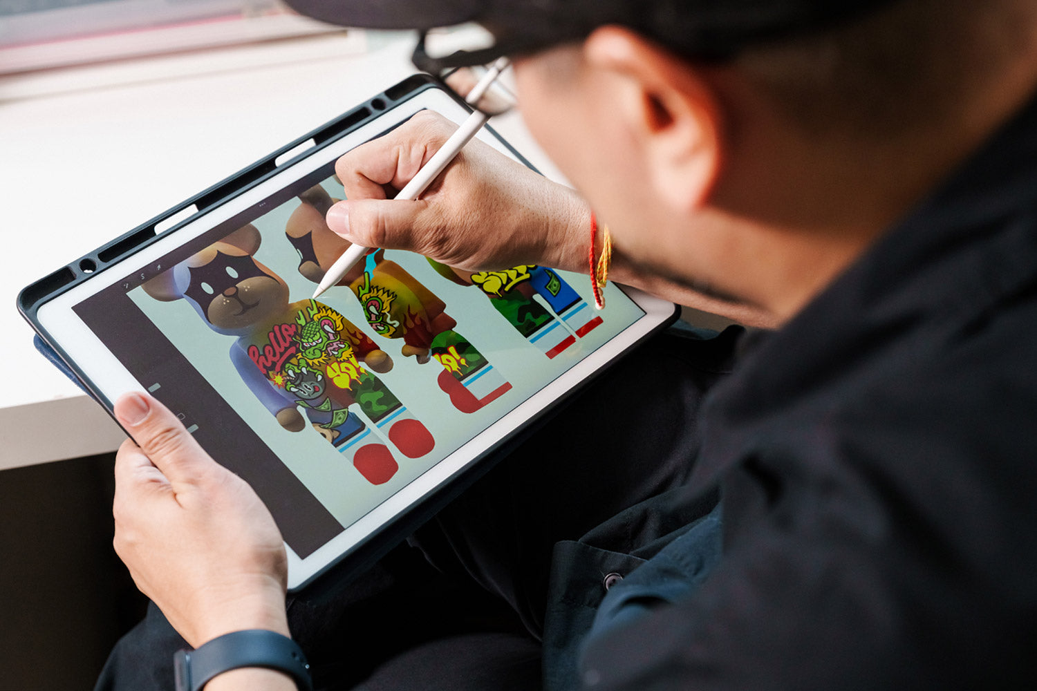 A person uses a stylus to digitally draw colorful designs on cartoon bear figures displayed on a tablet. The image is viewed from over the persons shoulder, focusing on their hand and the screen.