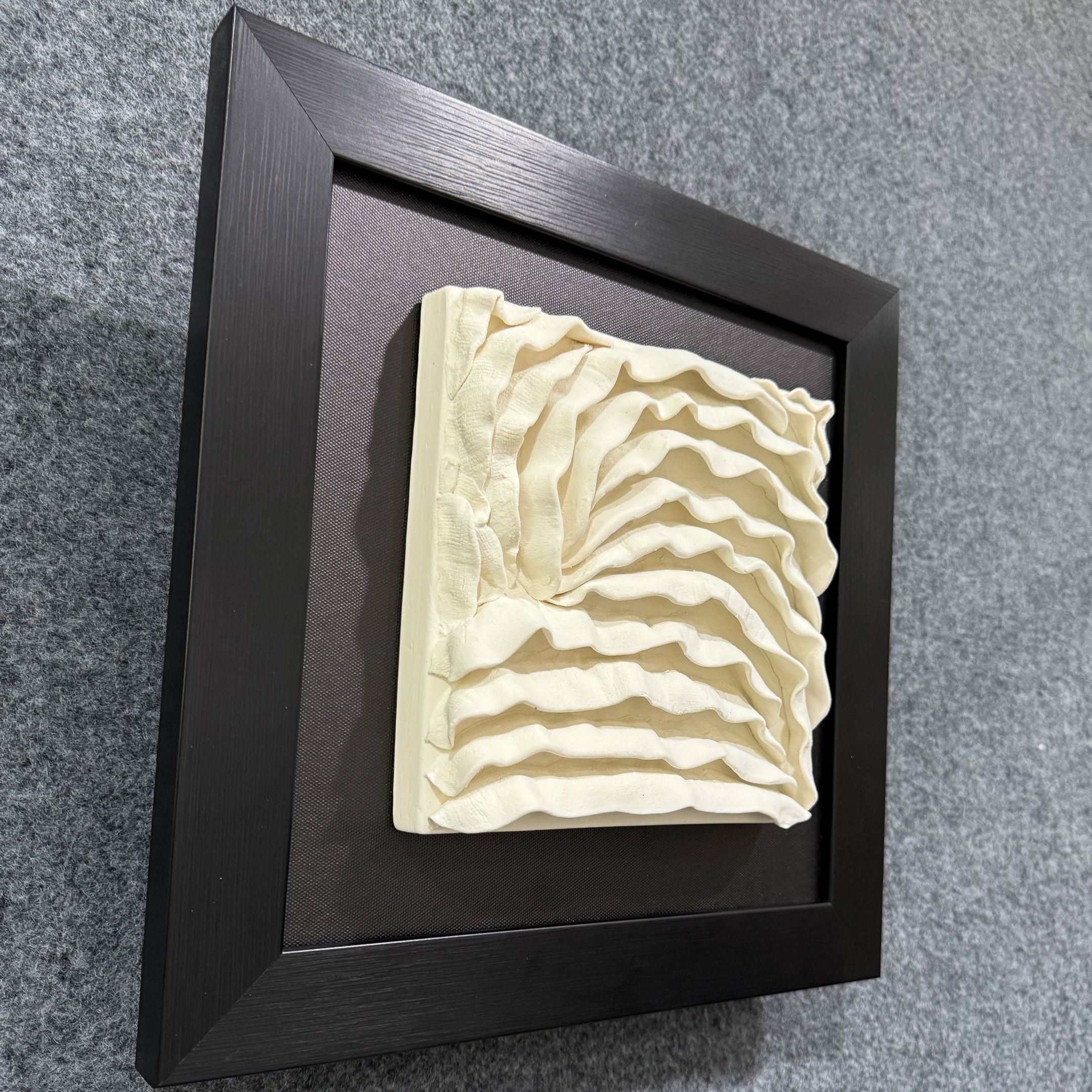 The Harmonia 3D Ceramic Wave Wall Sculpture by Giant Sculptures features layered cream ceramic waves in a textured, dimensional design set against a black frame, shown mounted on a gray carpeted surface.