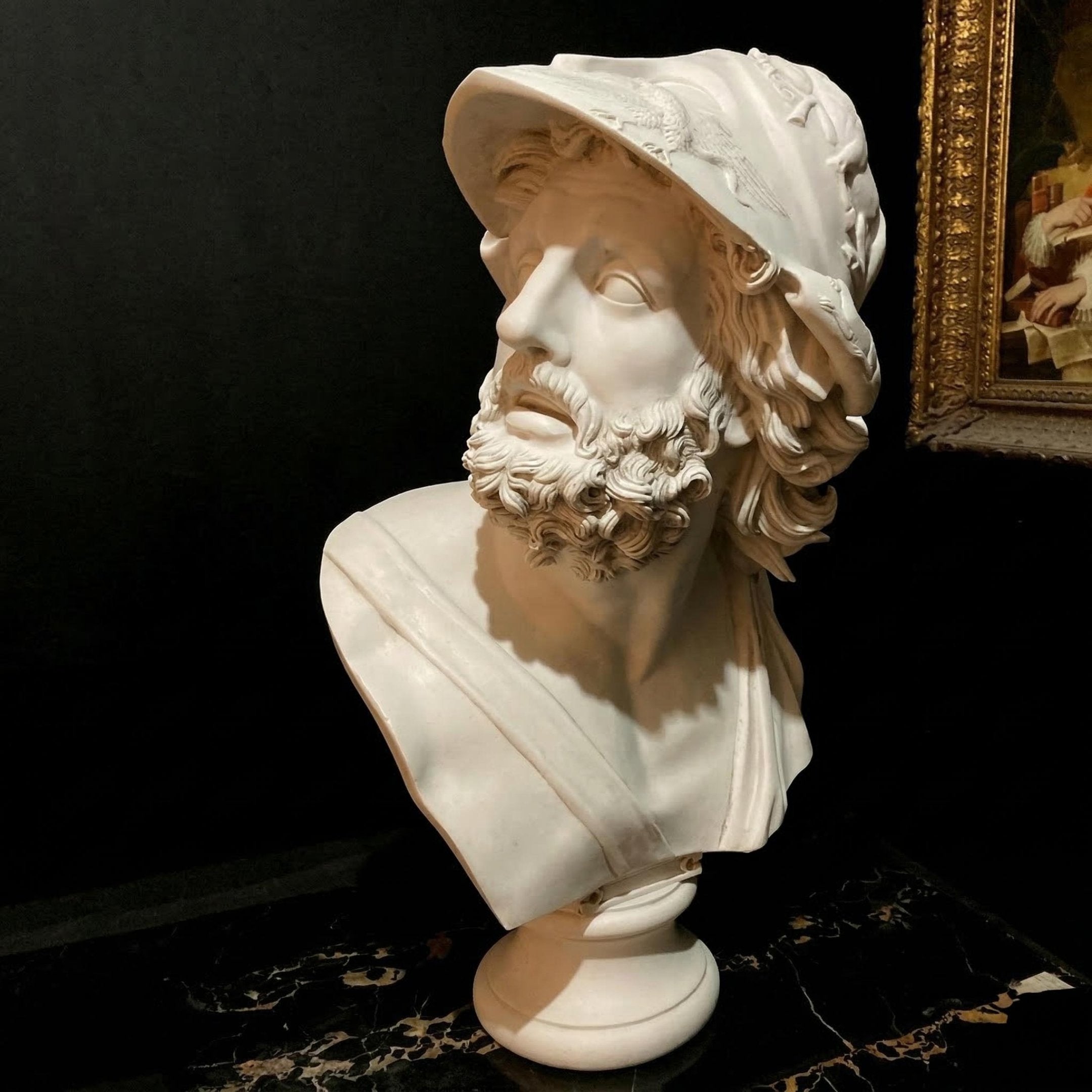 The Heroic Male Marble Bust - 65cm by Giant Sculptures features a bearded man in a decorated helmet, gazing left. Crafted in white marble, it stands on a dark surface with a gold-framed painting partly visible behind.