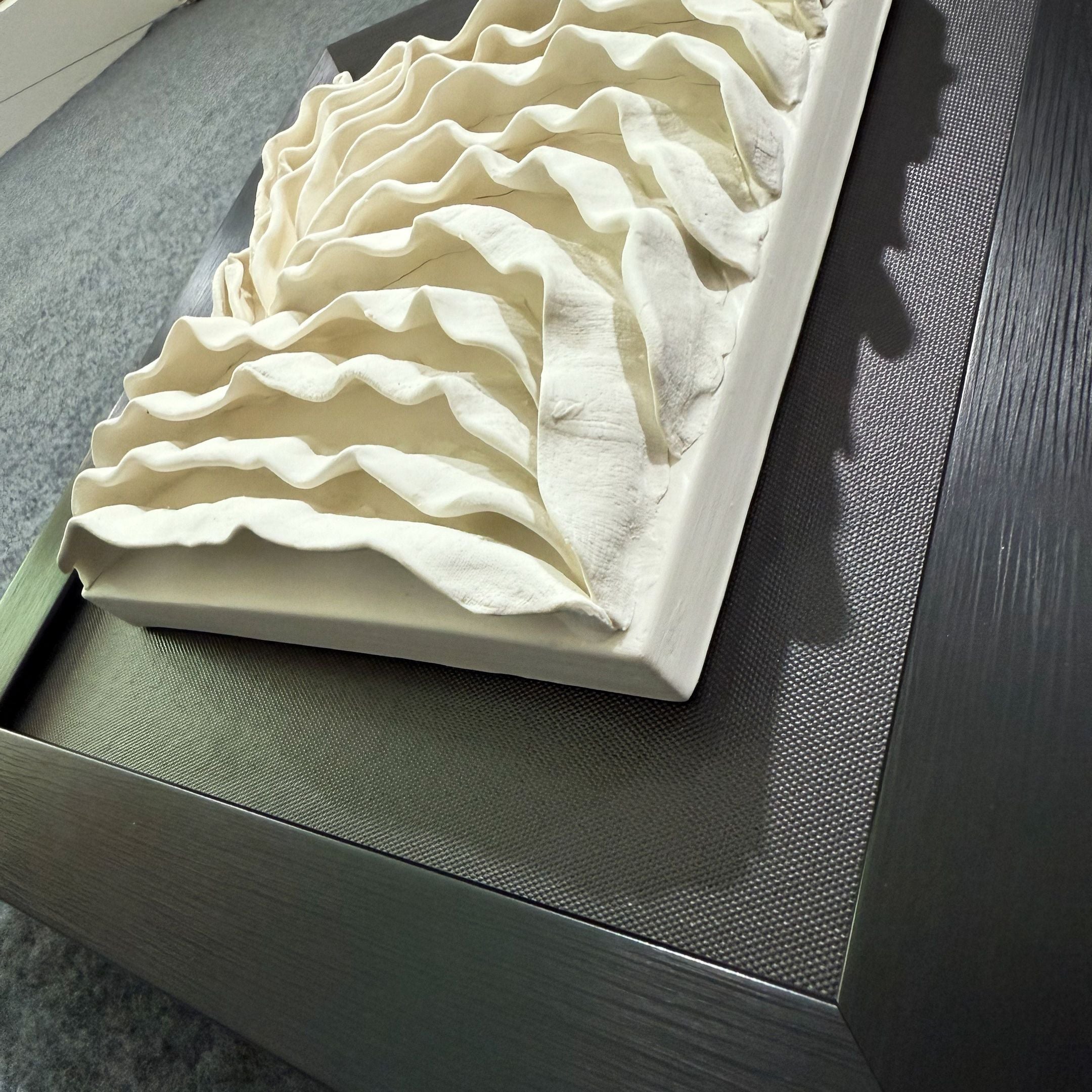 The Harmonia 3D Ceramic Wave Wall Sculpture by Giant Sculptures features wavy, layered white forms in a black frame, creating a modern art piece ideal for contemporary interiors.