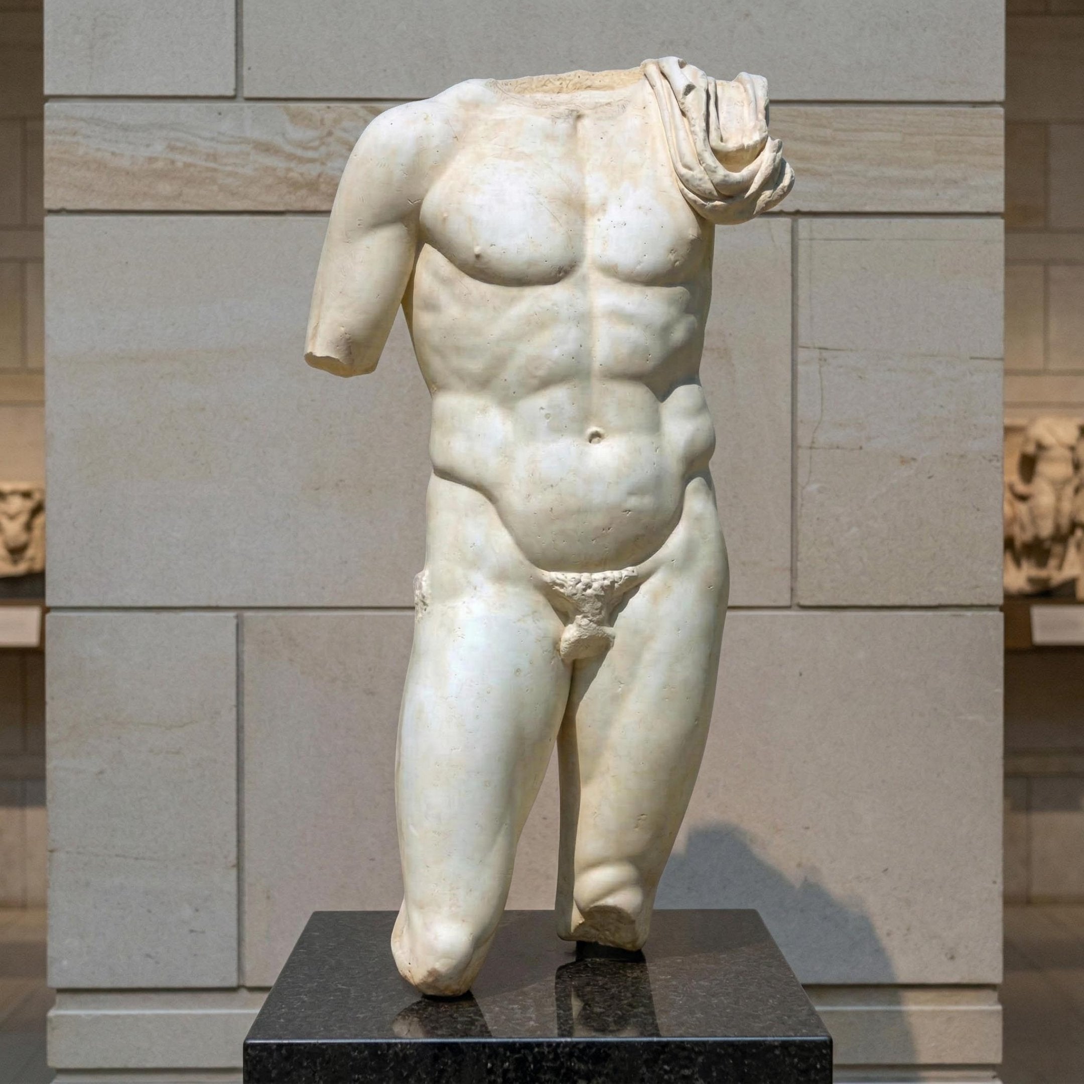 The Classical Male Marble Torso Sculpture - 130cm by Giant Sculptures features a muscular figure in natural white marble, draped over one shoulder and set on a black pedestal against a stone wall.