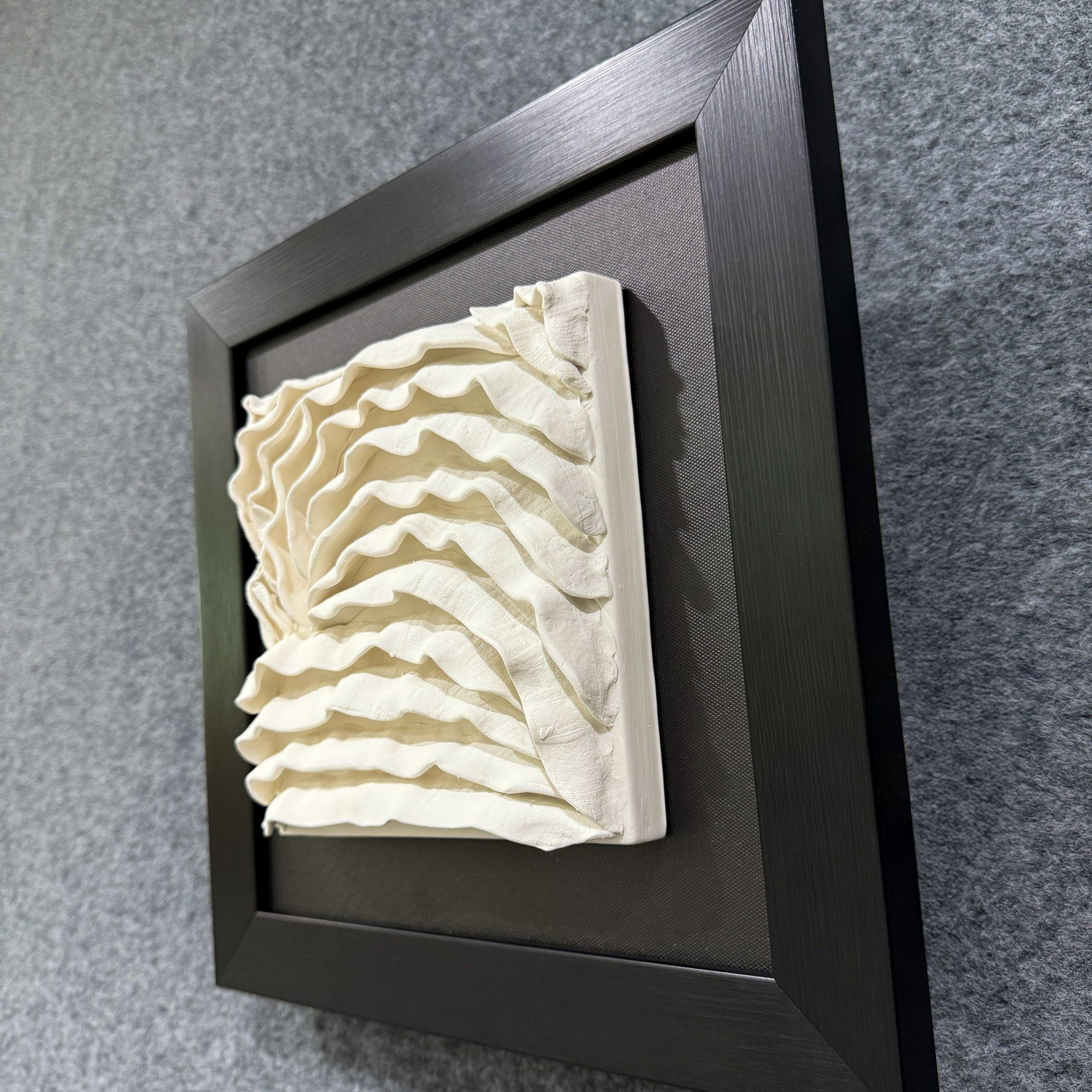 The Harmonia 3D Ceramic Wave Wall Sculpture With Black Frame by Giant Sculptures features layered, cream-colored ceramic waves in a striking three-dimensional pattern that enhances modern interiors against a dark backdrop.