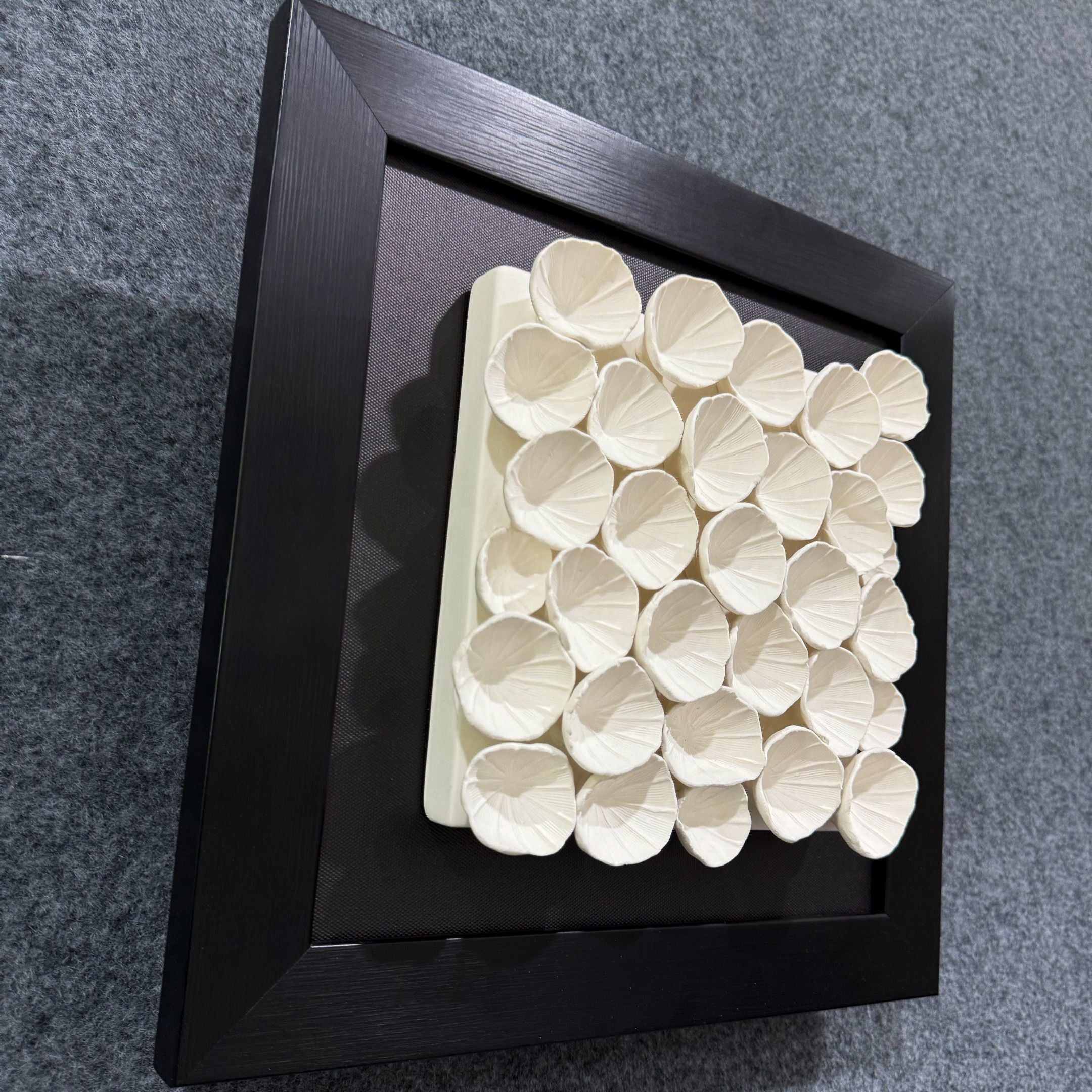 The Harmonia 3D Petal Wall Sculpture With Black Frame by Giant Sculptures features overlapping white ceramic petals in a grid on a raised rectangular base against a textured gray background—perfect for contemporary interiors.