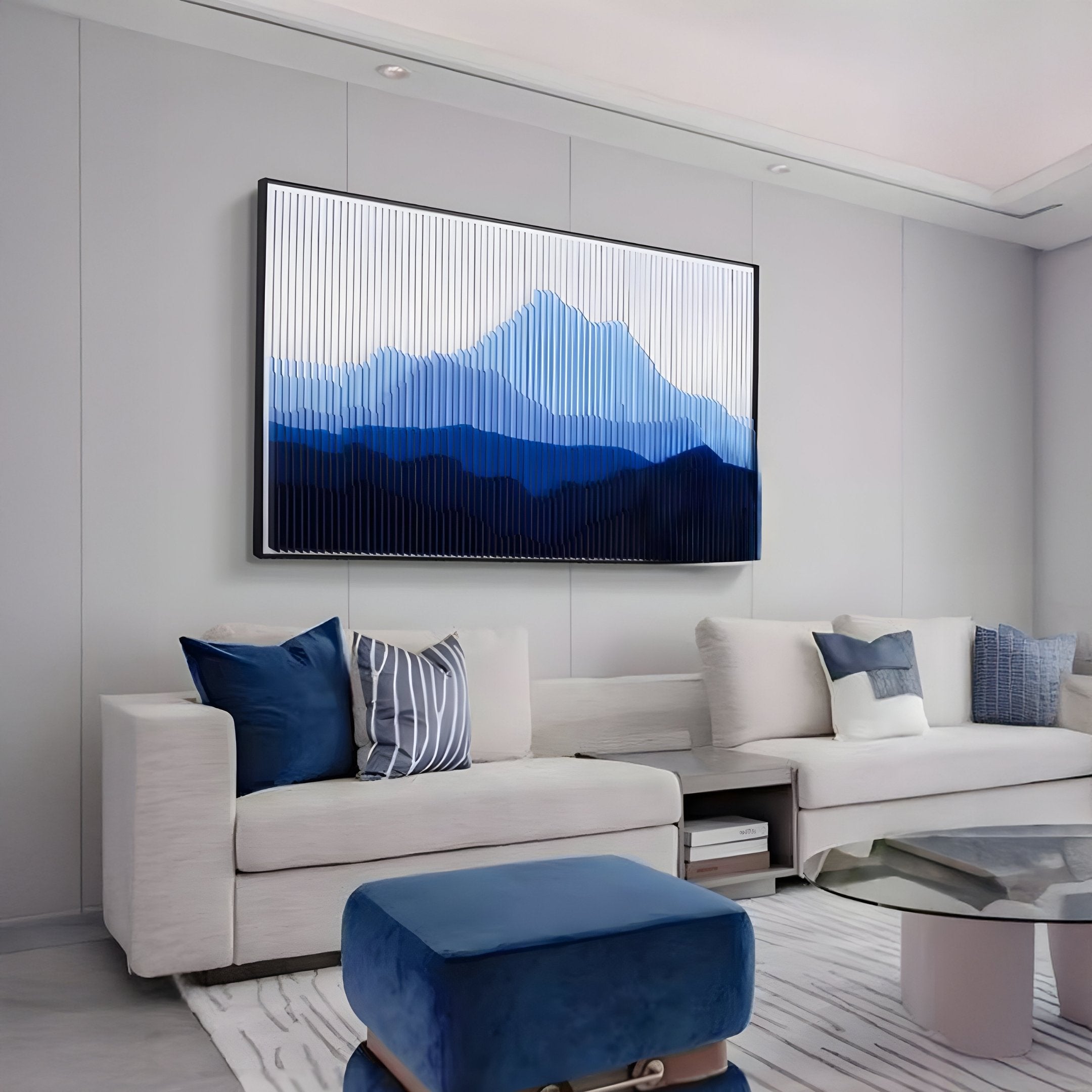 A modern living room with white sofas, blue and gray accent pillows, a glass coffee table, a blue ottoman, and Giant Sculptures’ Horizon Indigo Sky Mountain Acrylic 3D Wall Art as striking blue-toned wall décor.