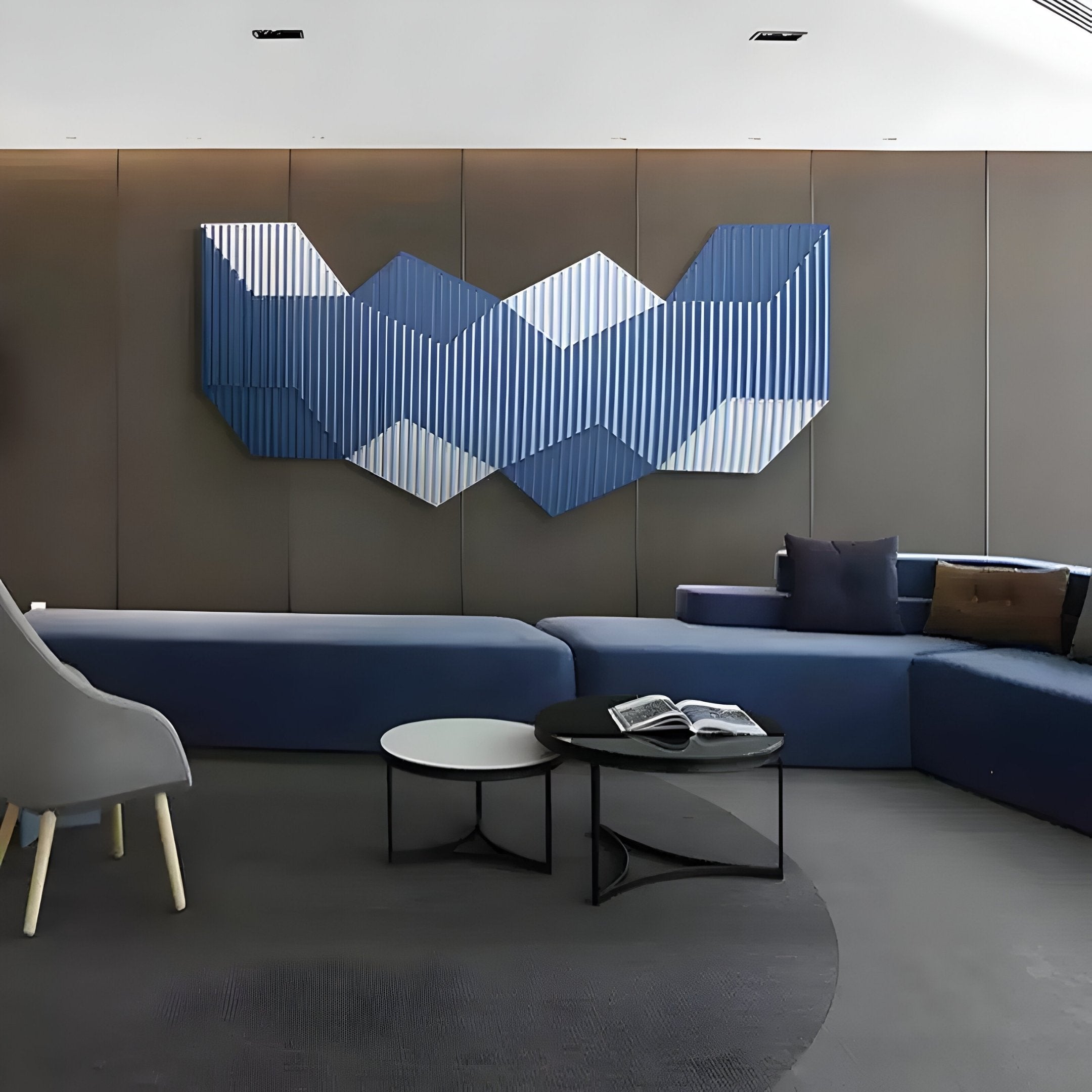 A modern lounge features a gray accent wall, Giant Sculptures Vertex Deep Navy & White Ridge Acrylic & Wood Carving 3D Wall Art, low blue seating, a gray armchair, two round nesting tables, and an open book for minimalist decor.