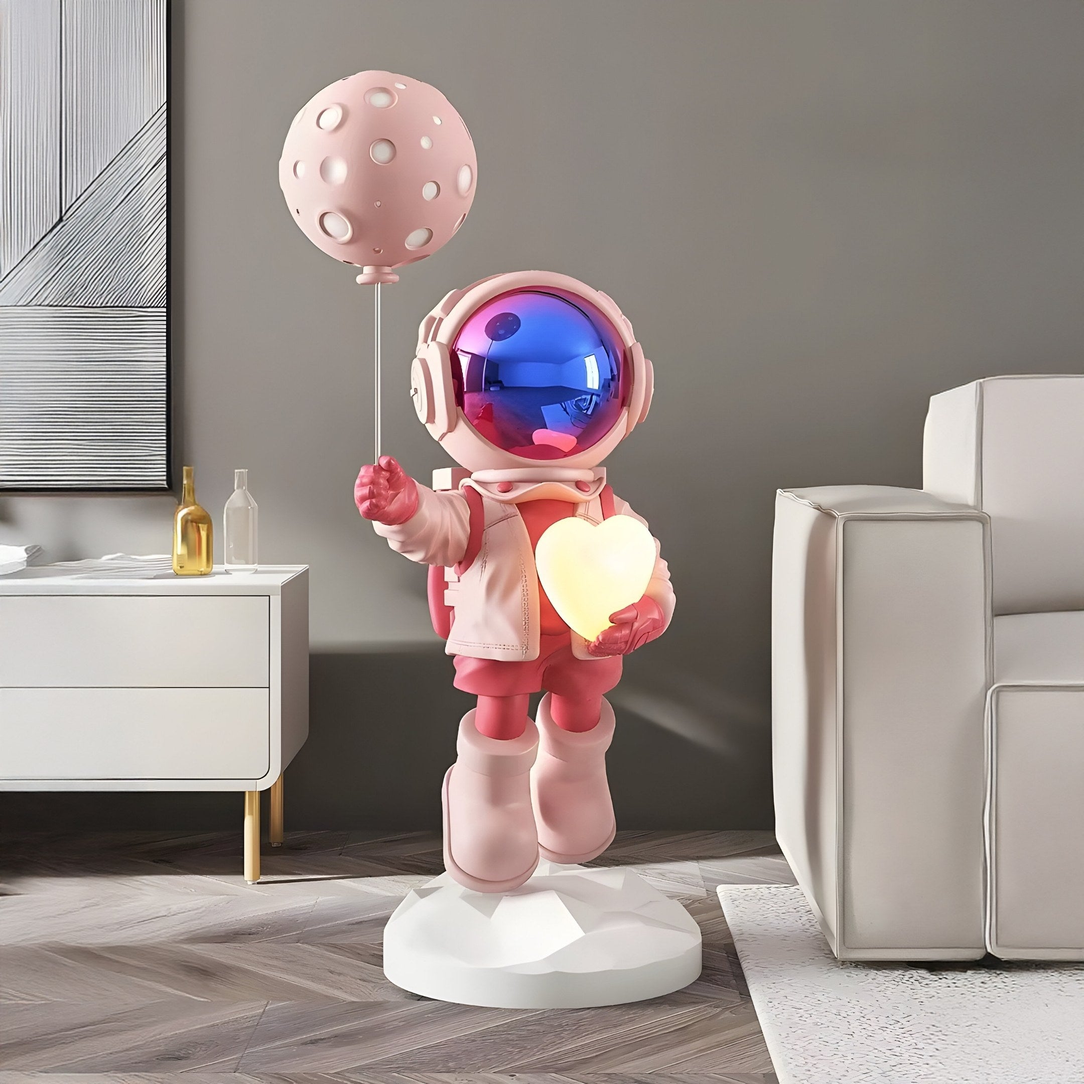 The Stellar Pink Astronaut Glow Heart Balloon Sculpture - 110cm by Giant Sculptures features a pink astronaut with a reflective blue visor, holding a glowing heart and pink balloon. It brings cosmic charm to any modern room, complemented by a white cabinet and gray sofa for an otherworldly yet stylish look.
