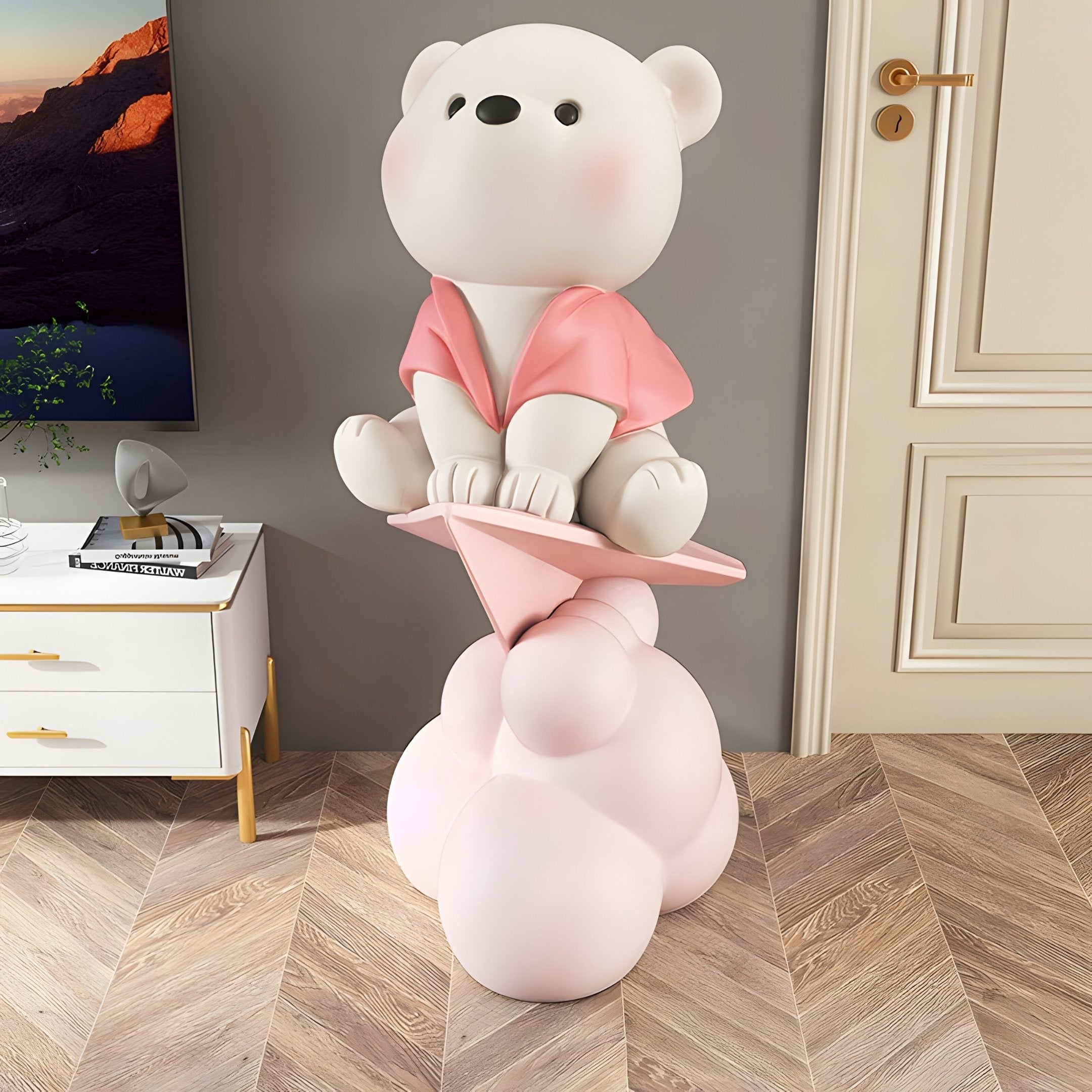 Blush Pink Cloudsit Glider Bear Sculpture by Giant Sculptures, a 150cm resin bear in ivory and soft pink seated on a paper glider atop layered pink cloud forms, shown in a living room beside a white cabinet.