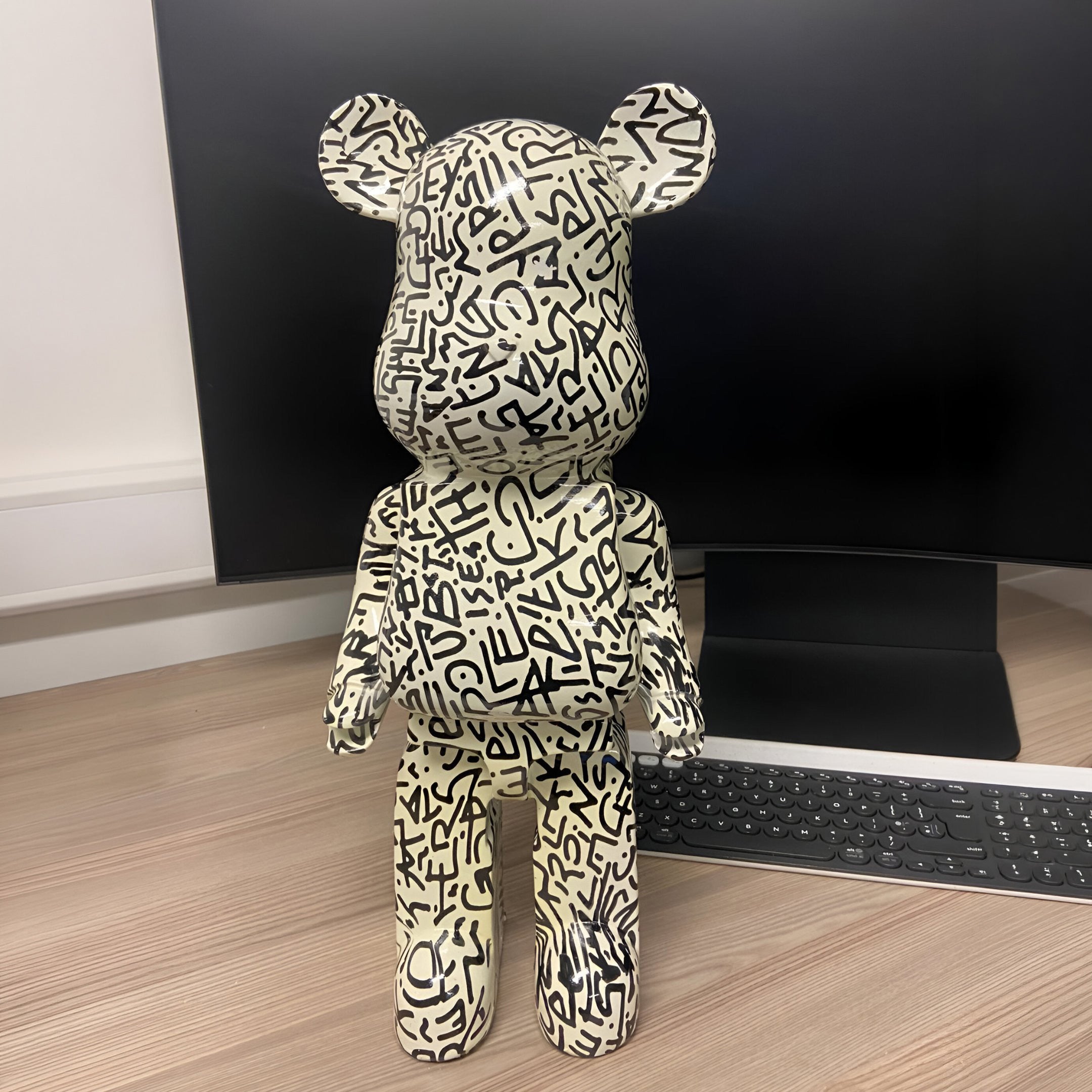 A 50cm Scribble Bear Sculpture by Giant Sculptures, featuring black graffiti-style writing, stands on a desk in front of a computer monitor and keyboard, facing away from the camera.