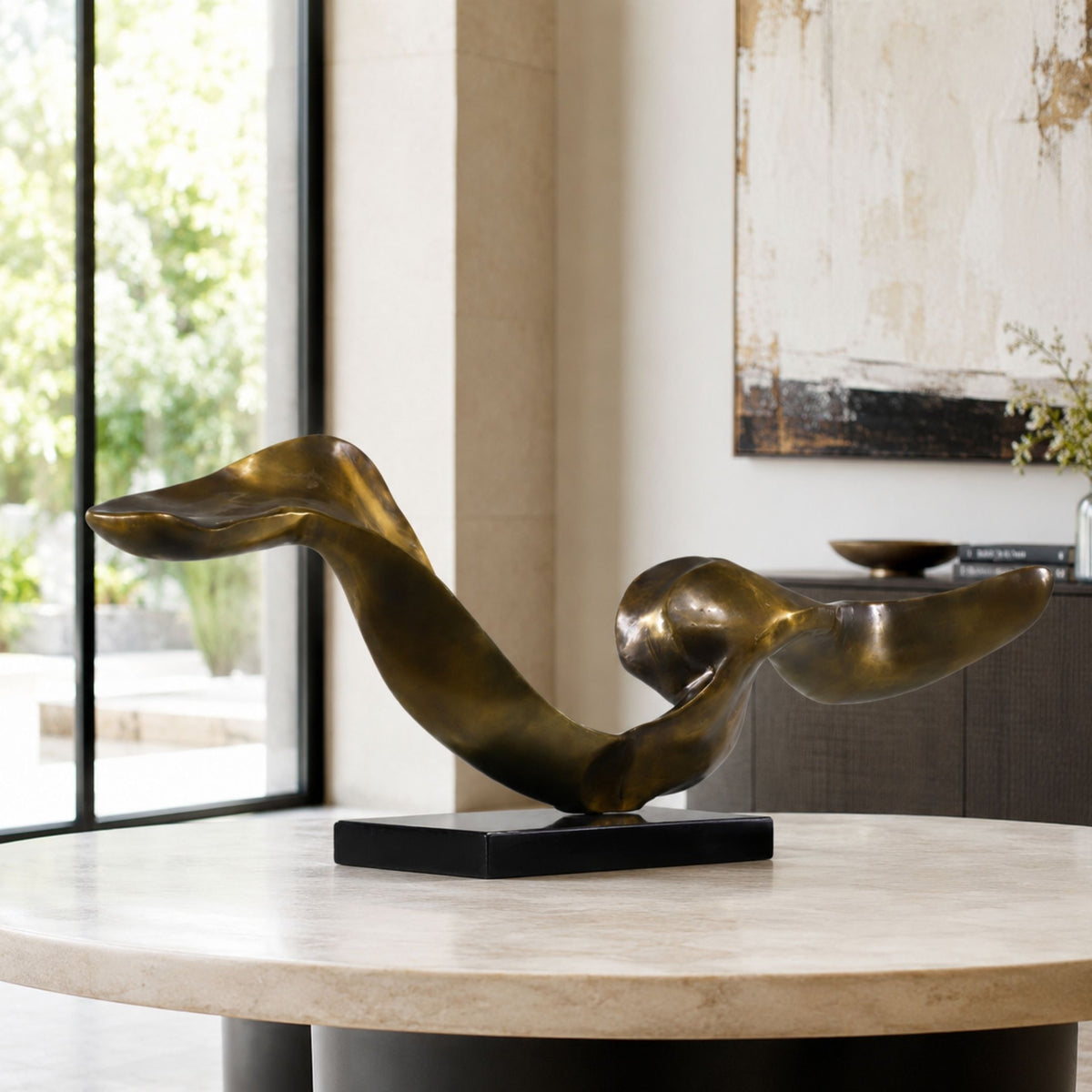 Bronze Abstract Flowing Ribbon Sculpture in brass on a black rectangular base, displayed on a round table in a living room near a window.