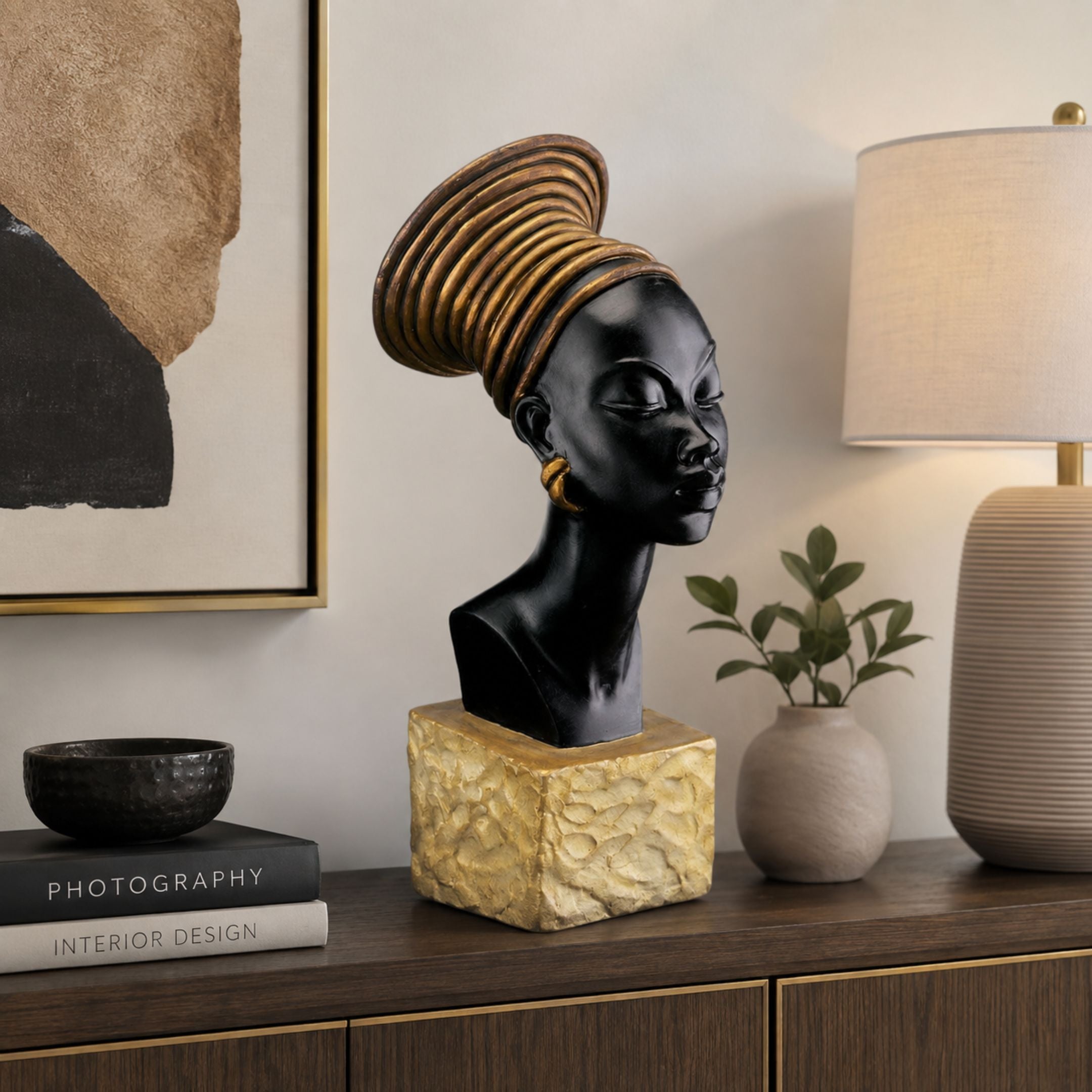 Contemporary African Female Bust Bronze Sculpture, 70cm, black finish with gold coiled headwrap on a gold textured base, shown on a console table beside a lamp and framed art.