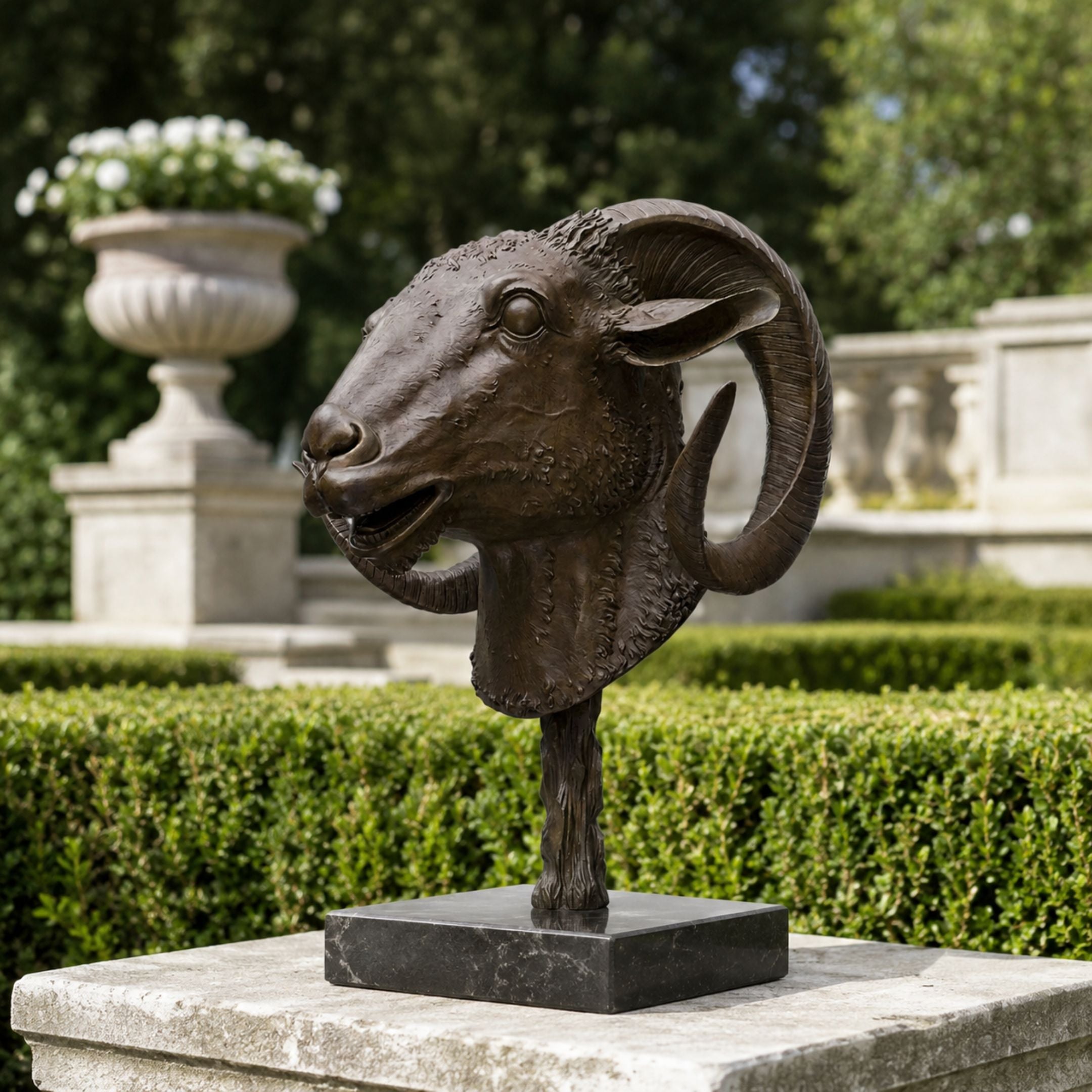 Traditional Ram Head Bronze Sculpture, 50cm, on a slender pole and dark marble base, displayed outdoors amid manicured hedges and stone garden décor.