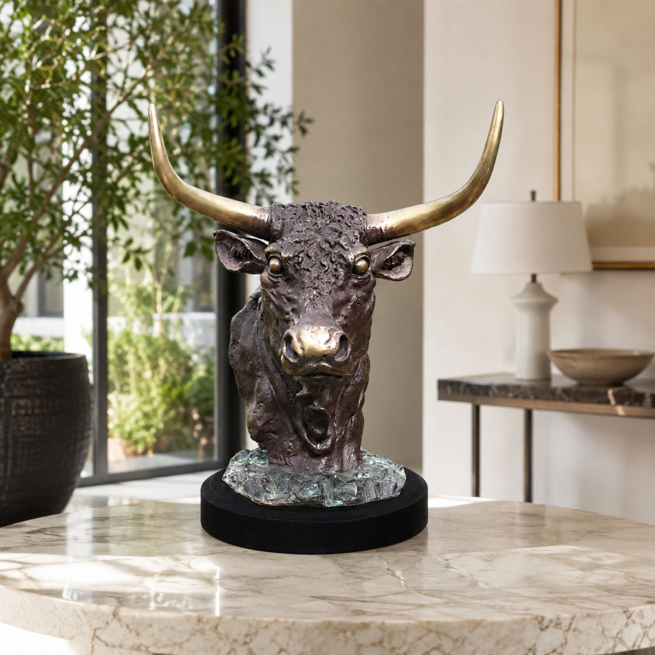 Traditional Bull Head Bronze Bust - 45cm displayed on a round black stand on a marble table, showing the longhorn bull's open mouth and gold-tipped horns in a living room setting.