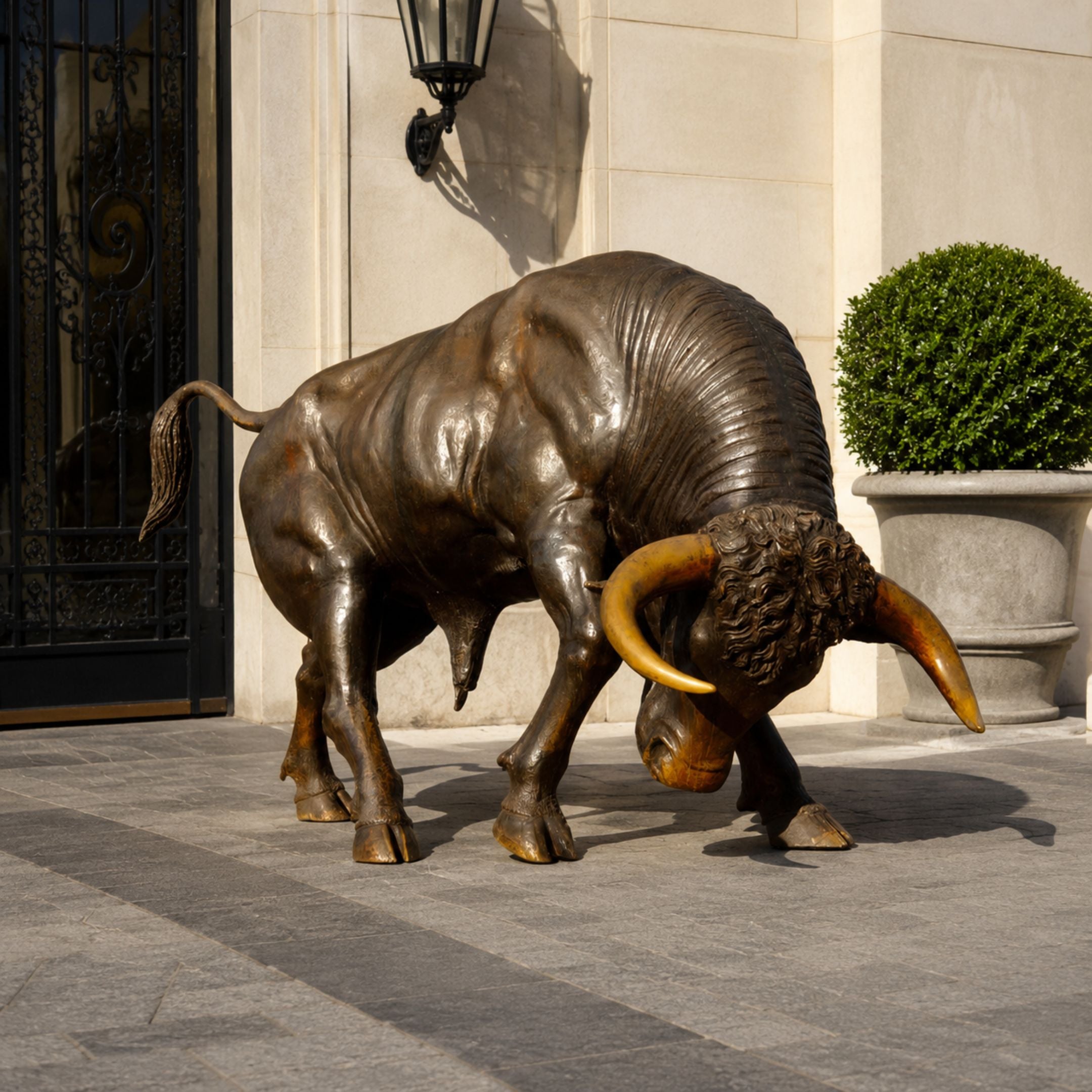 Large Contemporary Charging Bull Bronze Sculpture by Giant Sculptures with dark brown patina and golden horns, head lowered, displayed outside a stone building entrance.