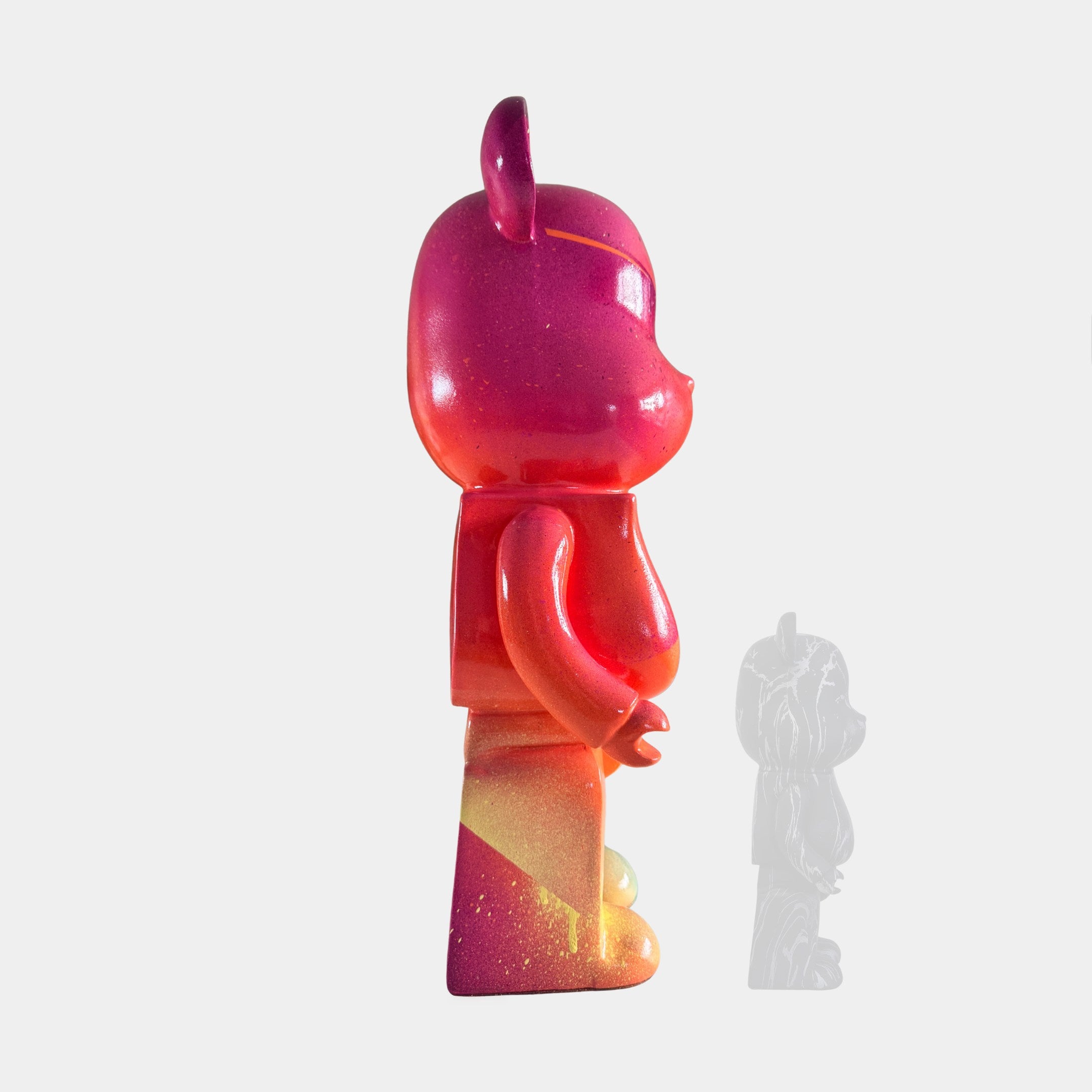 A side view of Giant Sculptures' Candy Bear Sculpture - 50cm, a colorful, limited edition art vinyl toy with pink, orange, and yellow gradients. It stands upright on a white background with a faint shadowy figure behind.