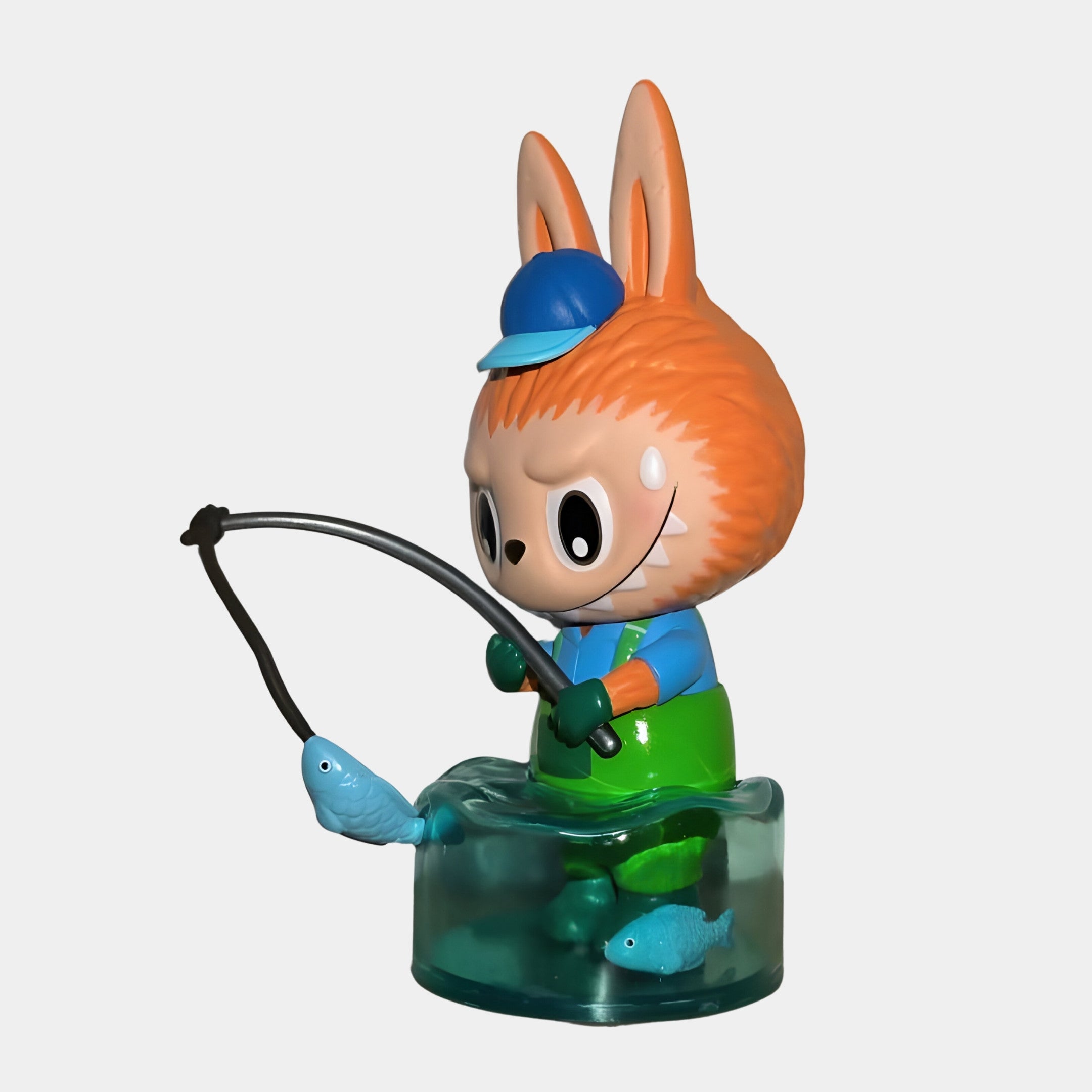 The Giant Sculptures Camp Life Fisher Labubu Sculpture - 100cm features a cute orange-furred character with big eyes and large ears, wearing a blue hat and shirt, holding a fish-tipped rod on a blue base with two blue fish nearby.