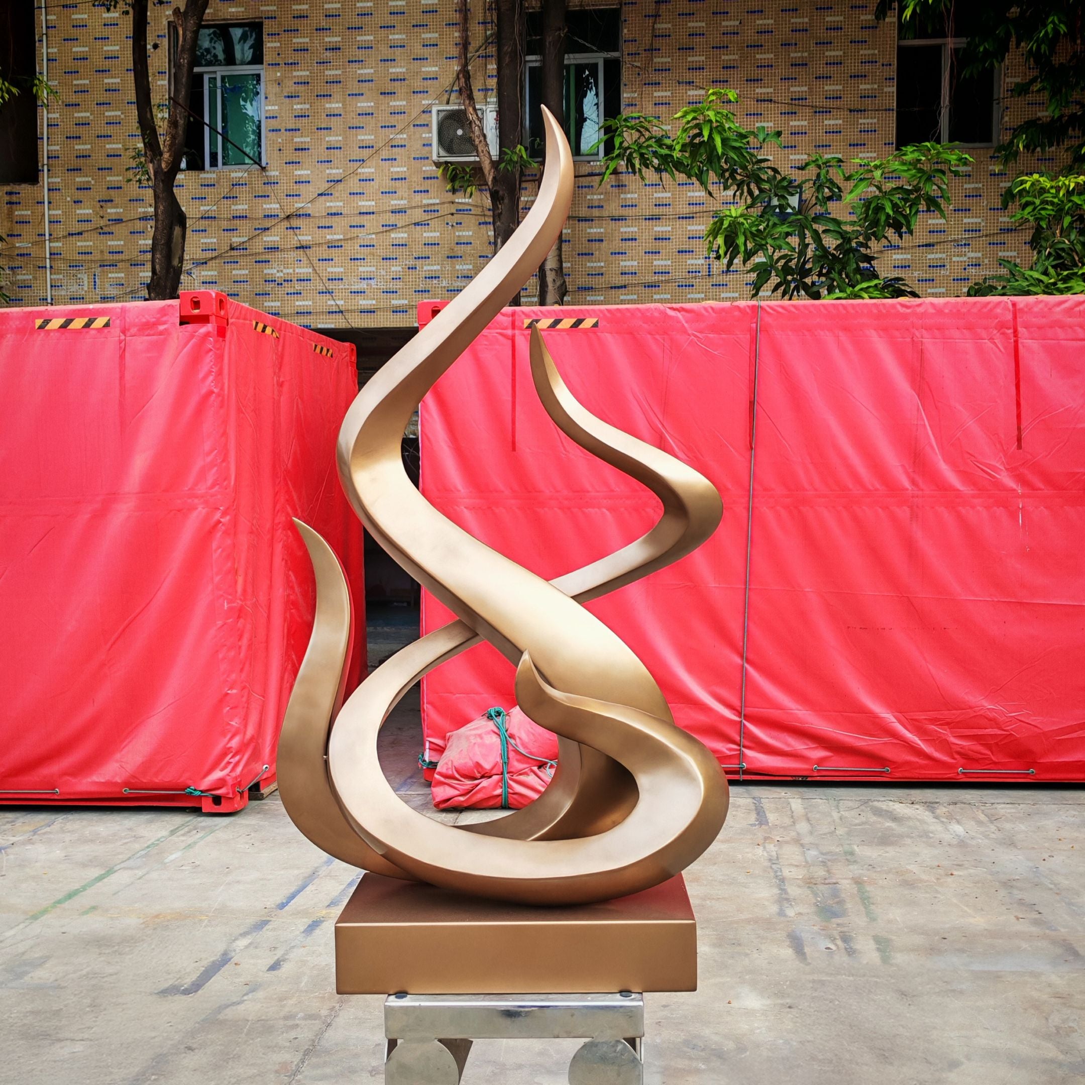 The Flame III Matt Bronze Abstract Steel Sculpture by Giant Sculptures features looping curves on a square base, displayed outdoors before two large red containers and a brick building with trees overhead.