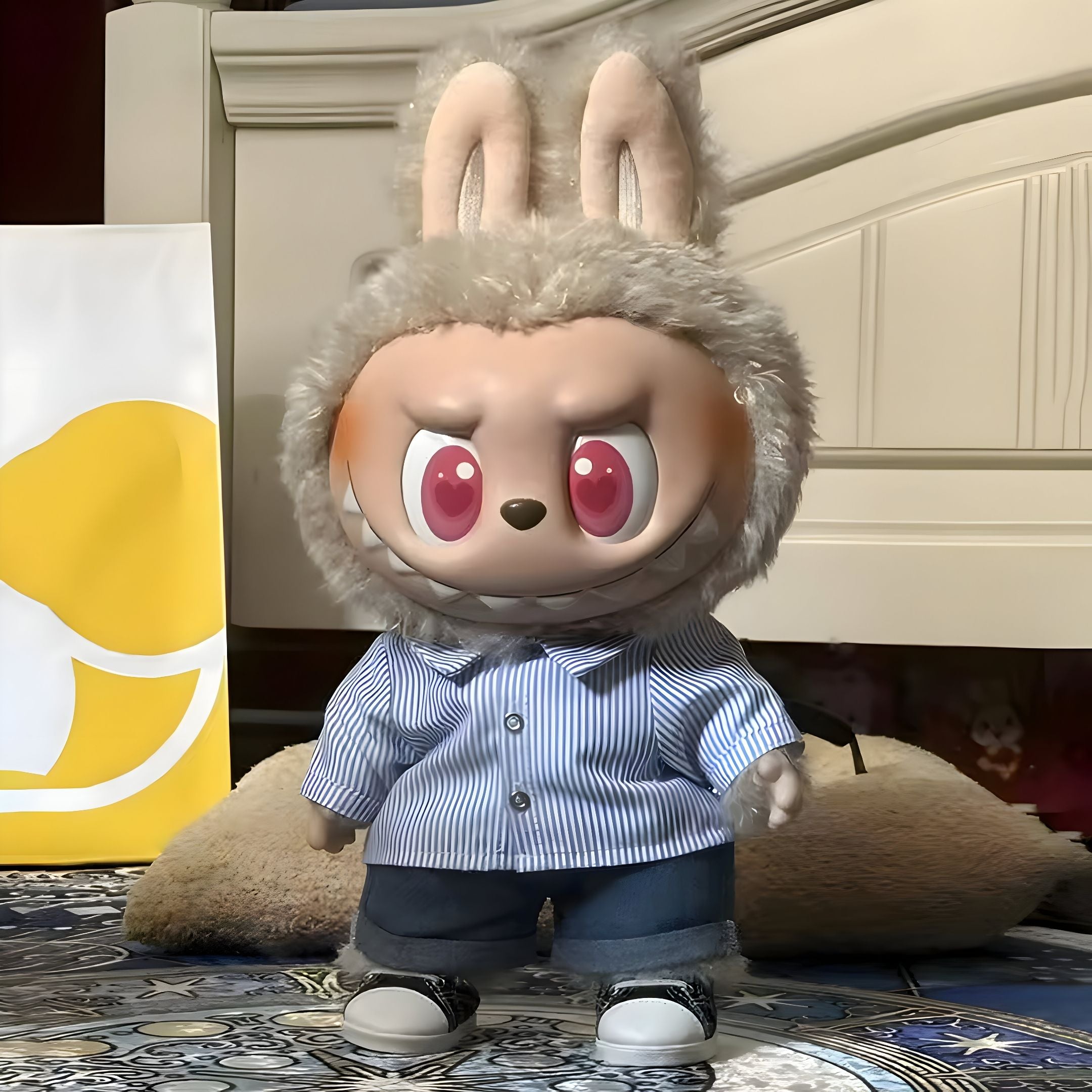 The Giant Sculptures Monster Flip With Me Labubu Sculpture - 100cm features big pink eyes, fluffy bunny ears, a mischievous grin, and wears a blue striped shirt, jeans, and sneakers. It stands on a patterned floor near a bed and shopping bag.