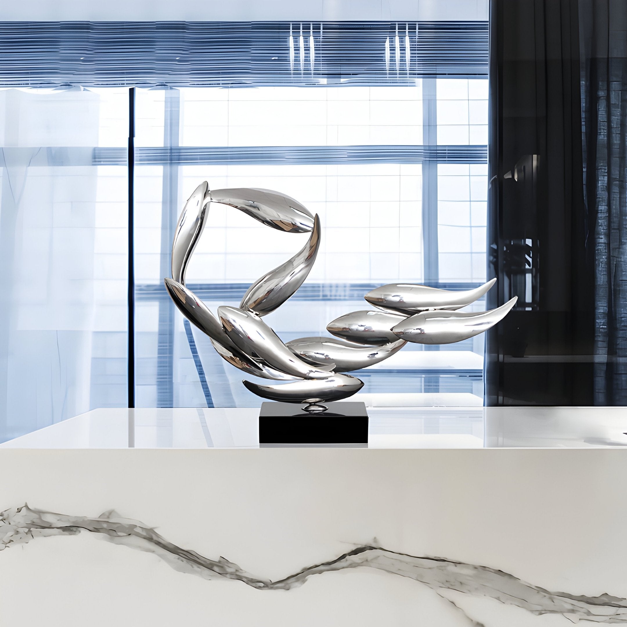 The Breeze Silver Organic Abstract Steel Sculpture by Giant Sculptures, showcasing modern, elongated, curved shapes, elegantly rests on a marble surface. Large windows with vertical blinds behind it softly diffuse light to highlight this captivating 50cm piece of art.
