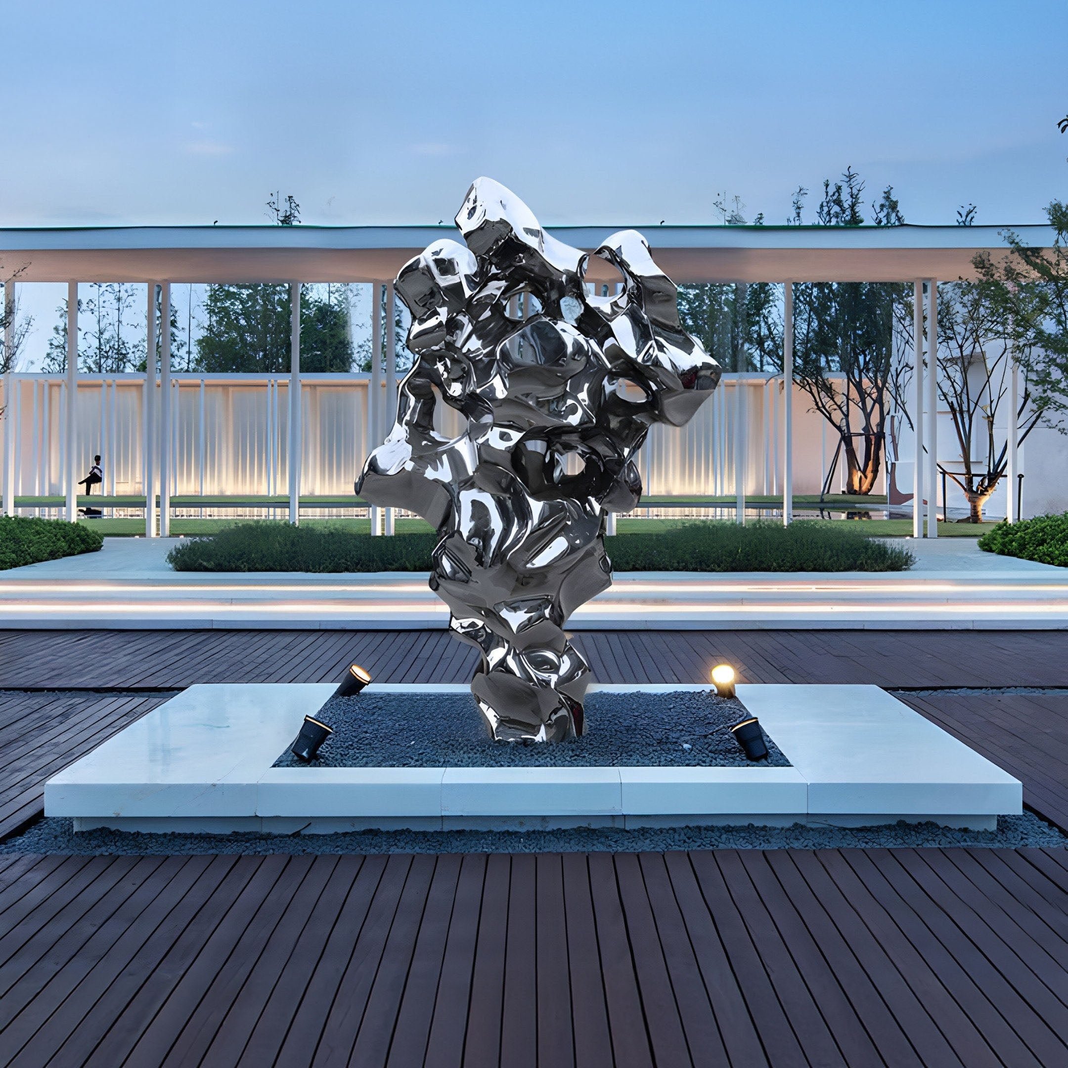 The Molten Silver Organic Steel Sculpture (155cm) by Giant Sculptures, an abstract stainless steel piece with a reflective surface, stands on a white platform outdoors. The backdrop features trees and a glass-walled building under clear skies, with pathways and outdoor lights enhancing the scene.