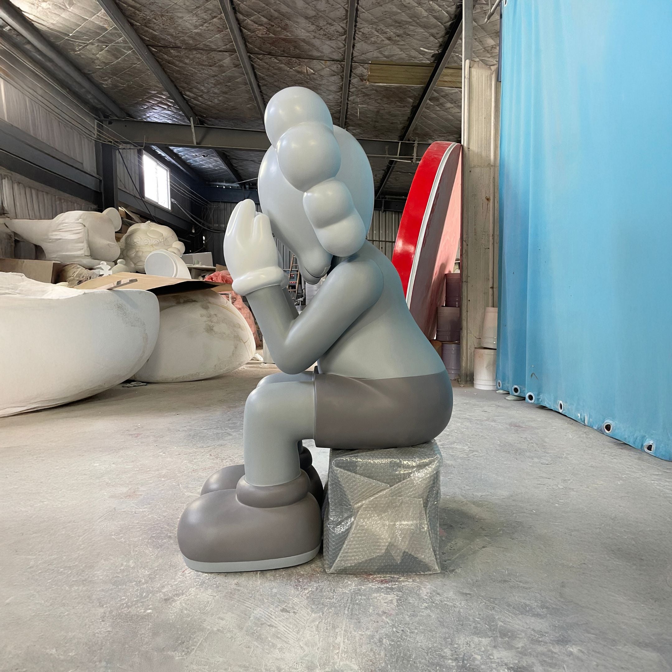 The Giant Sculptures Slate Grey Iconify Melancholy Sitting Sculpture (163cm) features a cartoon-like figure with gloved hands covering its face, seated on a metallic box in an industrial warehouse with materials and equipment in the background.