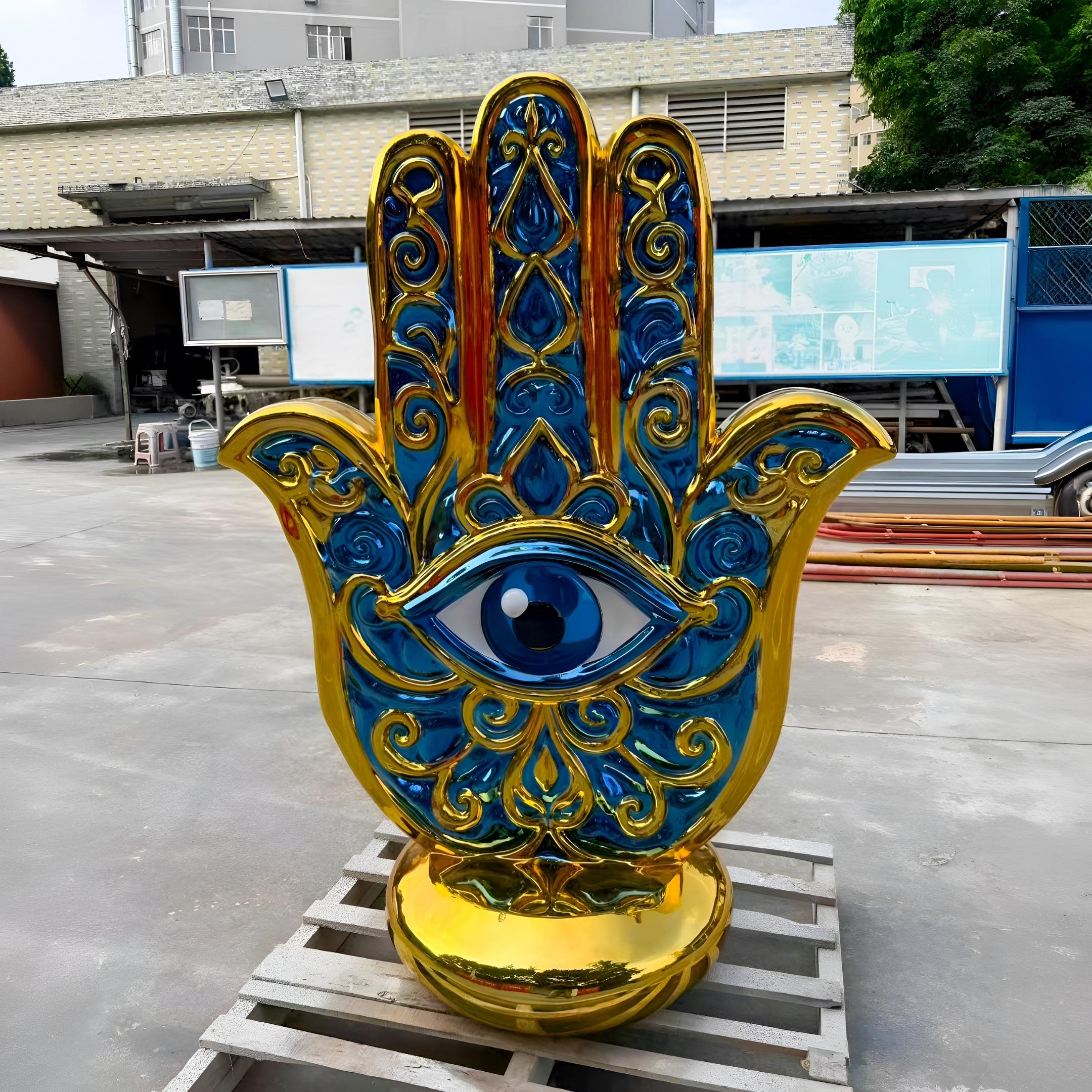 Giant Sculptures' 150cm Golden Hamsa Hand Protection Sculpture in cobalt blue and gold fibreglass with central eye motif, displayed on a pallet in an outdoor workshop yard.
