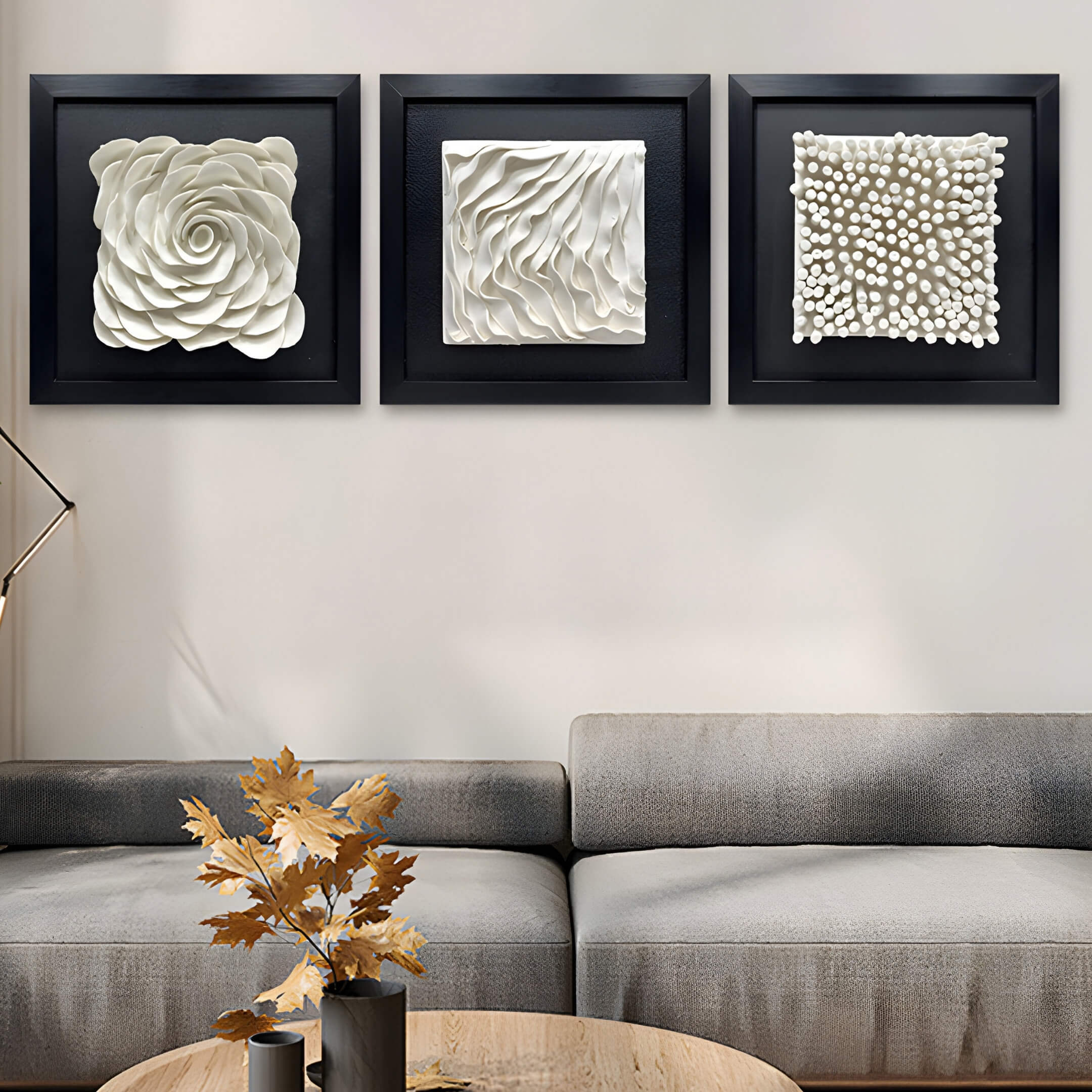 Above the beige sofa, three square, black-framed Harmonia 3D Rose Wall Installations from Giant Sculptures feature white textured patterns: rose-like swirls, wavy lines, and circular bumps. A small table with a vase of dried leaves completes the modern interior décor.