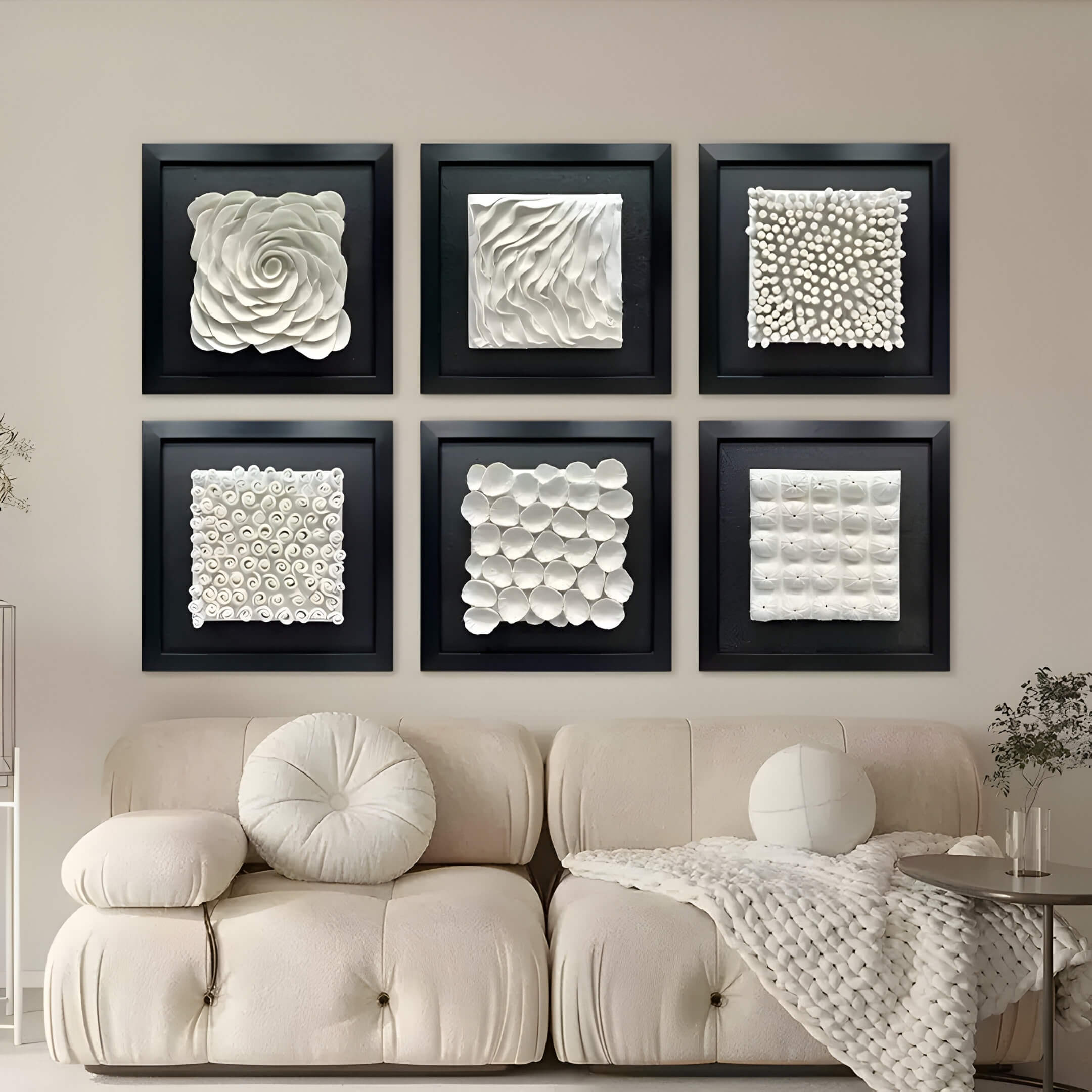 A cozy living room features a cream couch with round and square pillows. Above, six Harmonia 3D Spiral Wall Sculptures by Giant Sculptures in a 2x3 grid, boast textured white designs framed in black to enhance the modern aesthetic. A small plant adds natural elegance to the space.