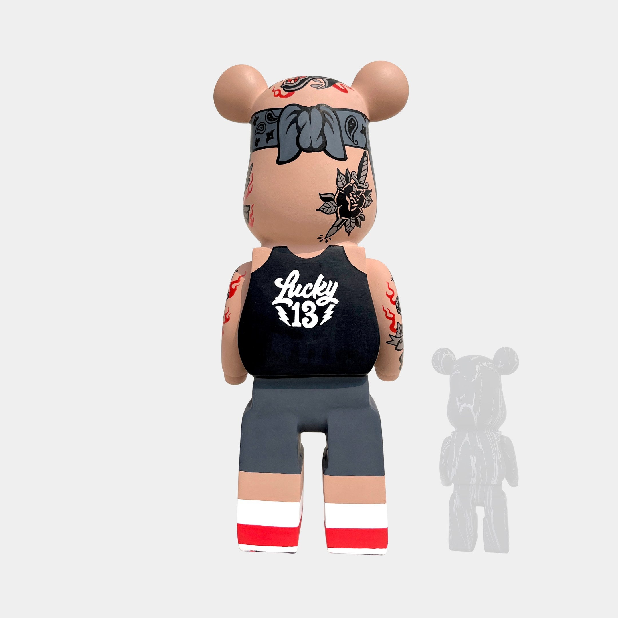 The Iron Paw Bear Sculpture - 135cm by Giant Sculptures features a bear-shaped vinyl form with tattoos, a black Lucky 13 tank top, and gray shorts, standing on a white background with its shadow to the right.