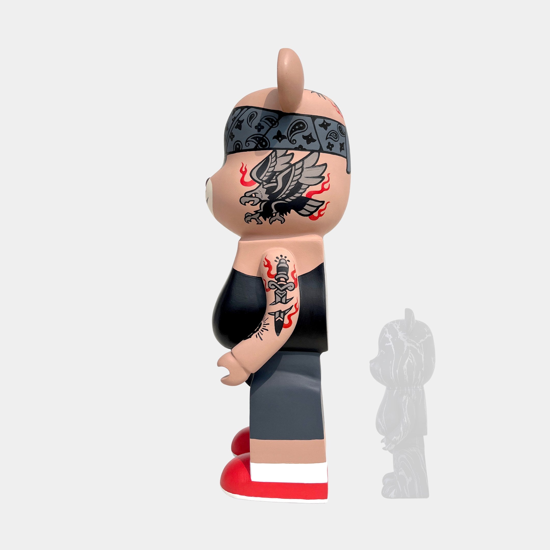 A side view of the Giant Sculptures Iron Paw Bear Sculpture - 135cm limited edition, a bear-shaped vinyl toy inspired by street artist BigDel, featuring tattoos, black tank top, gray shorts, red and white shoes, and a patterned headband.