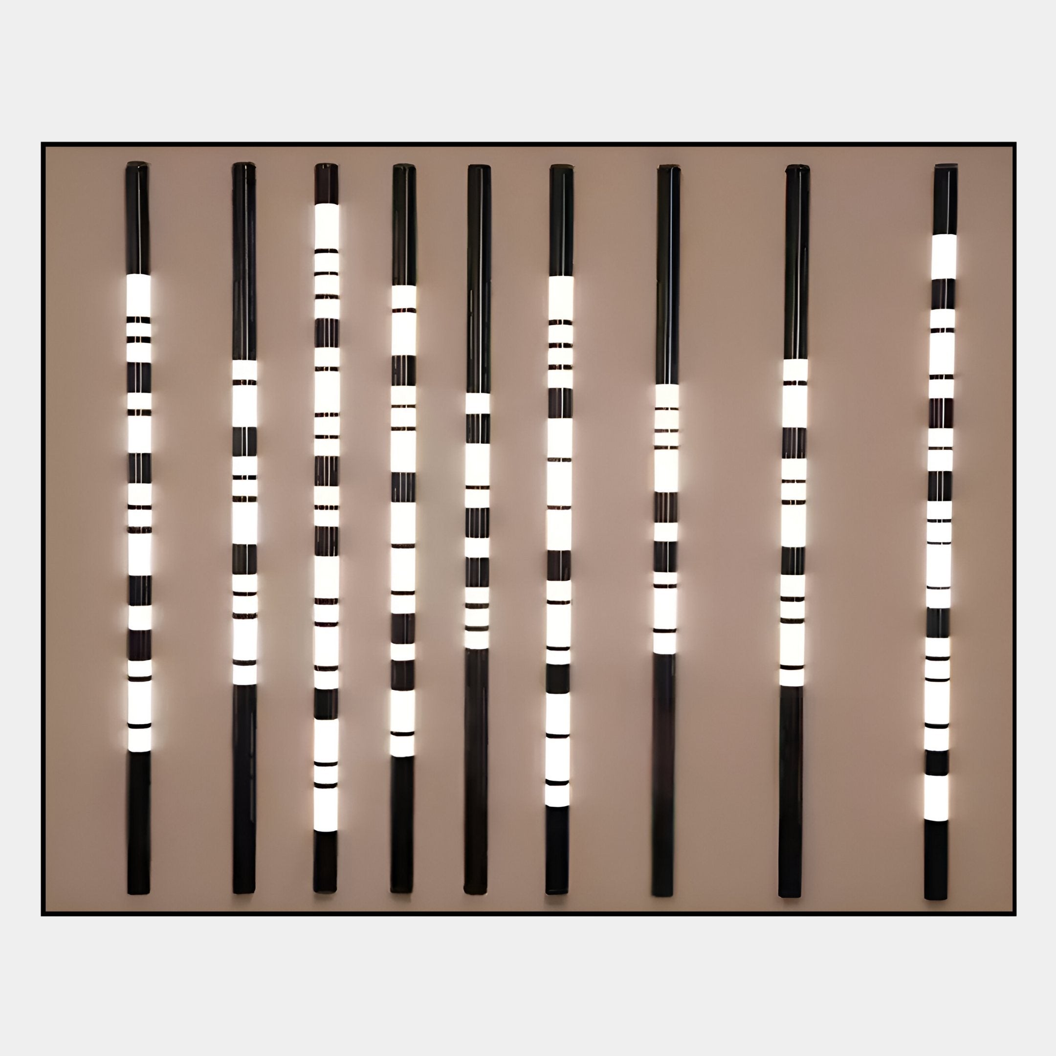 The Strata Taupe LED Wall Art by Giant Sculptures features nine vertical black rods with illuminated white bands in varying widths and positions, creating a geometric, minimalist design against a taupe wall—ideal for modern interiors.