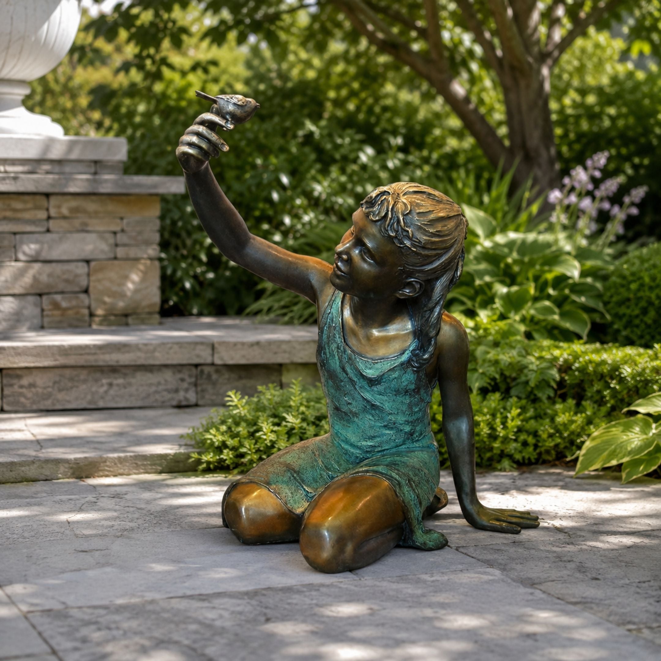 Large Contemporary Girl with Bird Bronze Sculpture, 130cm, kneeling girl in patinated bronze raising a bird skyward, displayed on a stone patio among garden greenery.