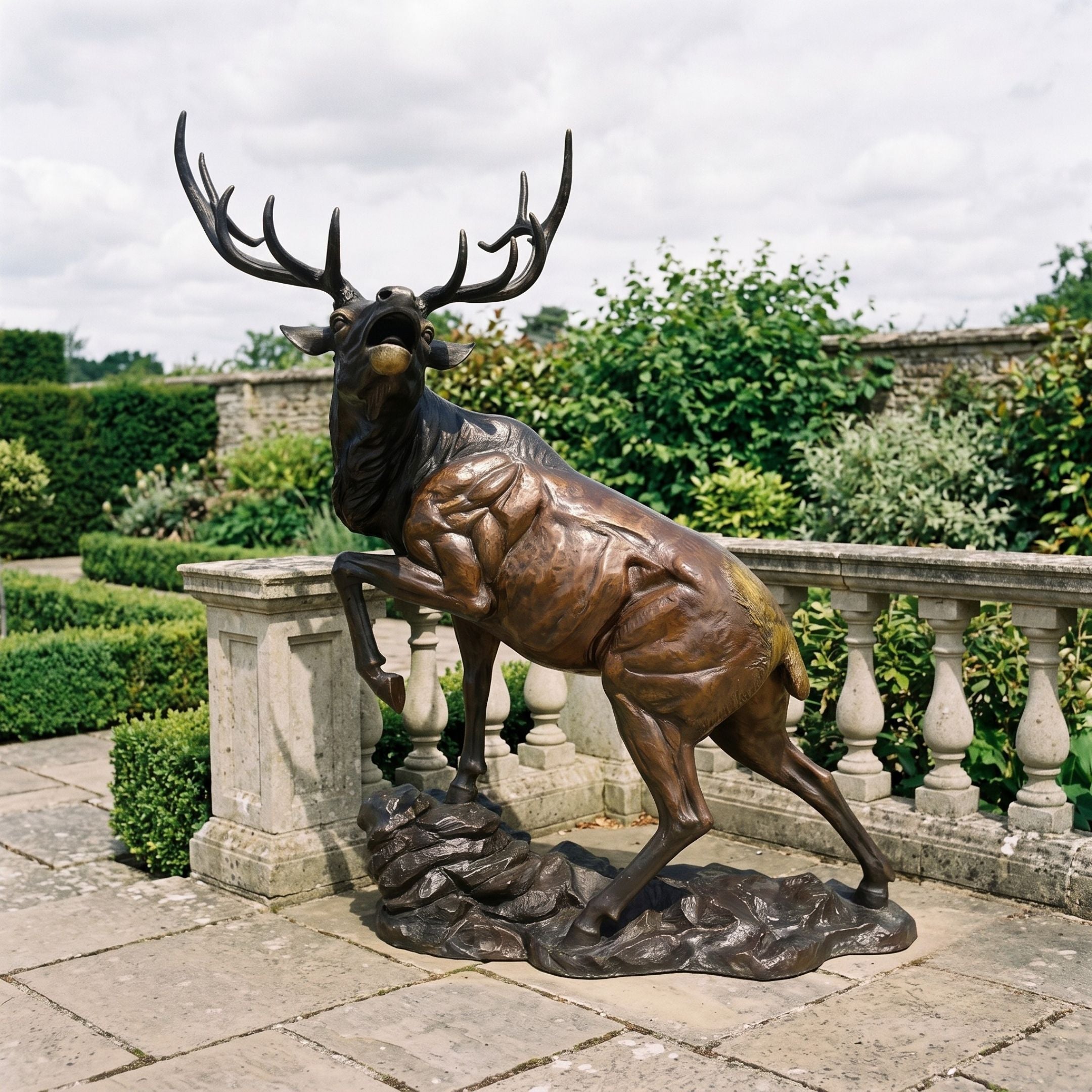 Life-Size Contemporary Bugling Elk Bronze Sculpture by Giant Sculptures in warm amber and dark patina, head raised mid-bugle on a rocky base beside a garden balustrade.