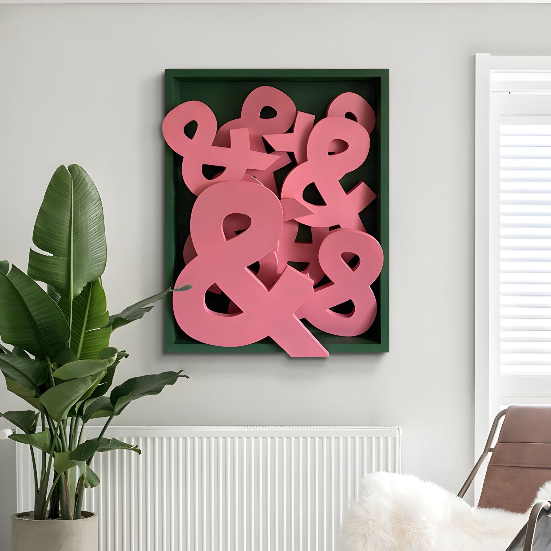 The Typova Pink Ampersand Wood Carving 3D Wall Art by Giant Sculptures showcases large pink ampersands overlapping on a dark green background, displayed above a white radiator beside a leafy plant in a bright, modern room.