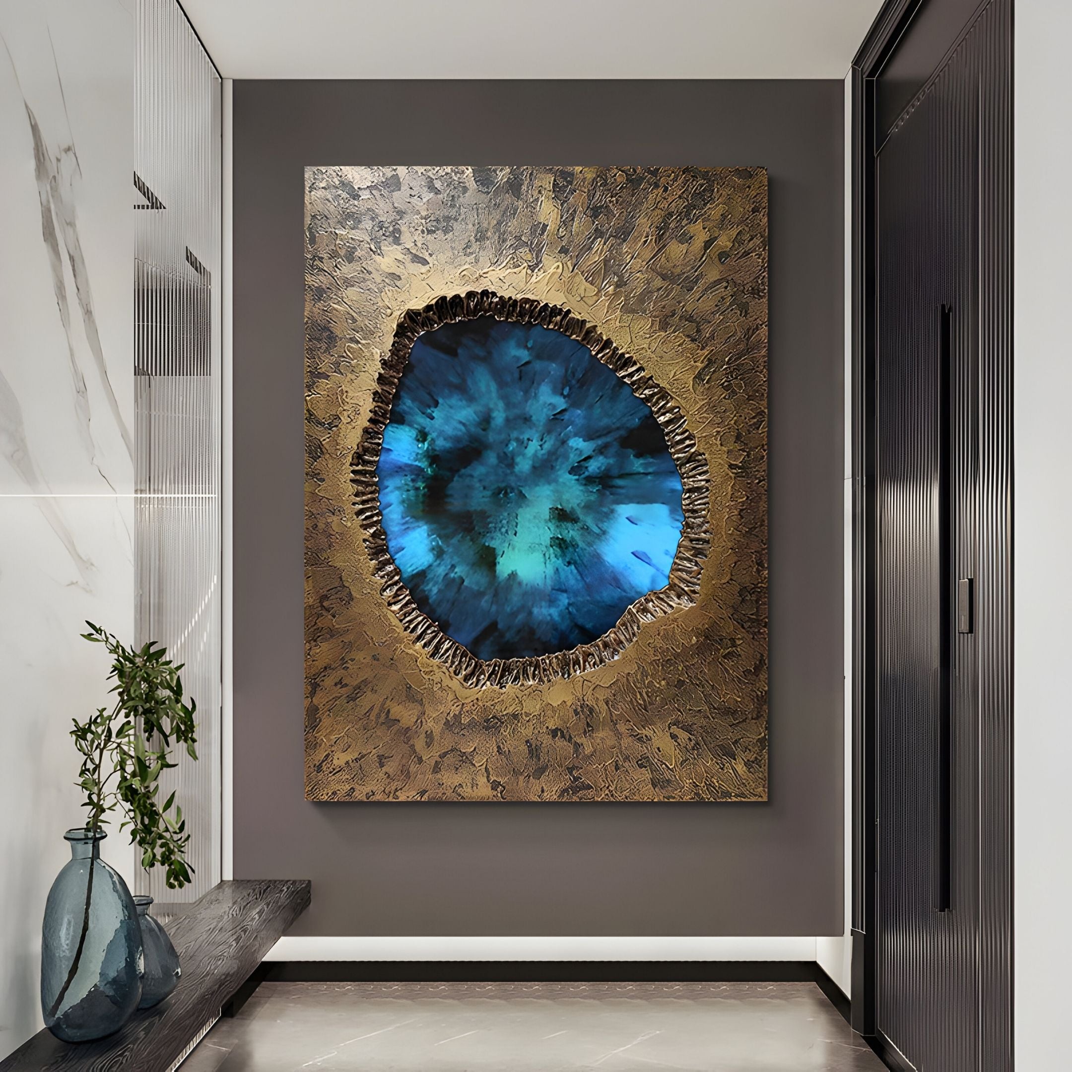 The Vorthera Sapphire Burst Textured Gold 3D Wall Art by Giant Sculptures with a blue geode-like center and gold edges hangs on a gray wall above a glass vase with greenery in a modern hallway accented by marble and dark wood.