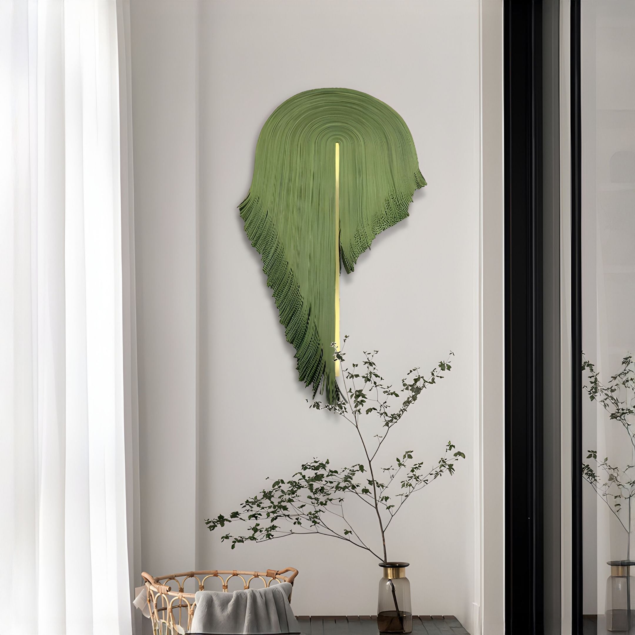 The Arclune Olive Drift Sculptural Felt 3D Wall Art by Giant Sculptures, resembling a palm leaf, hangs above a tall glass vase with branches in a modern room with sheer white curtains and abundant natural light.