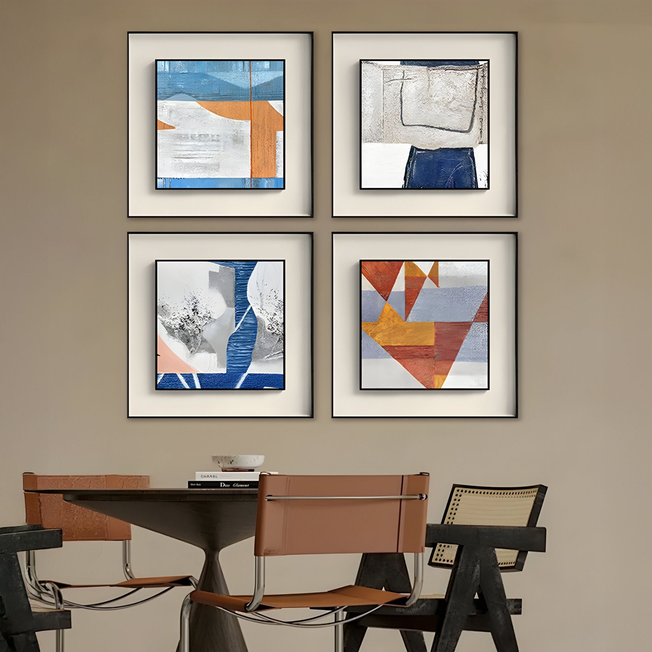A modern dining area with two tan chairs at a round table sits beneath the Aerolith Blue & Ivory Abstract Textured Framed Wall Art by Giant Sculptures, featuring four blue, orange, and gray pieces on the beige wall above.